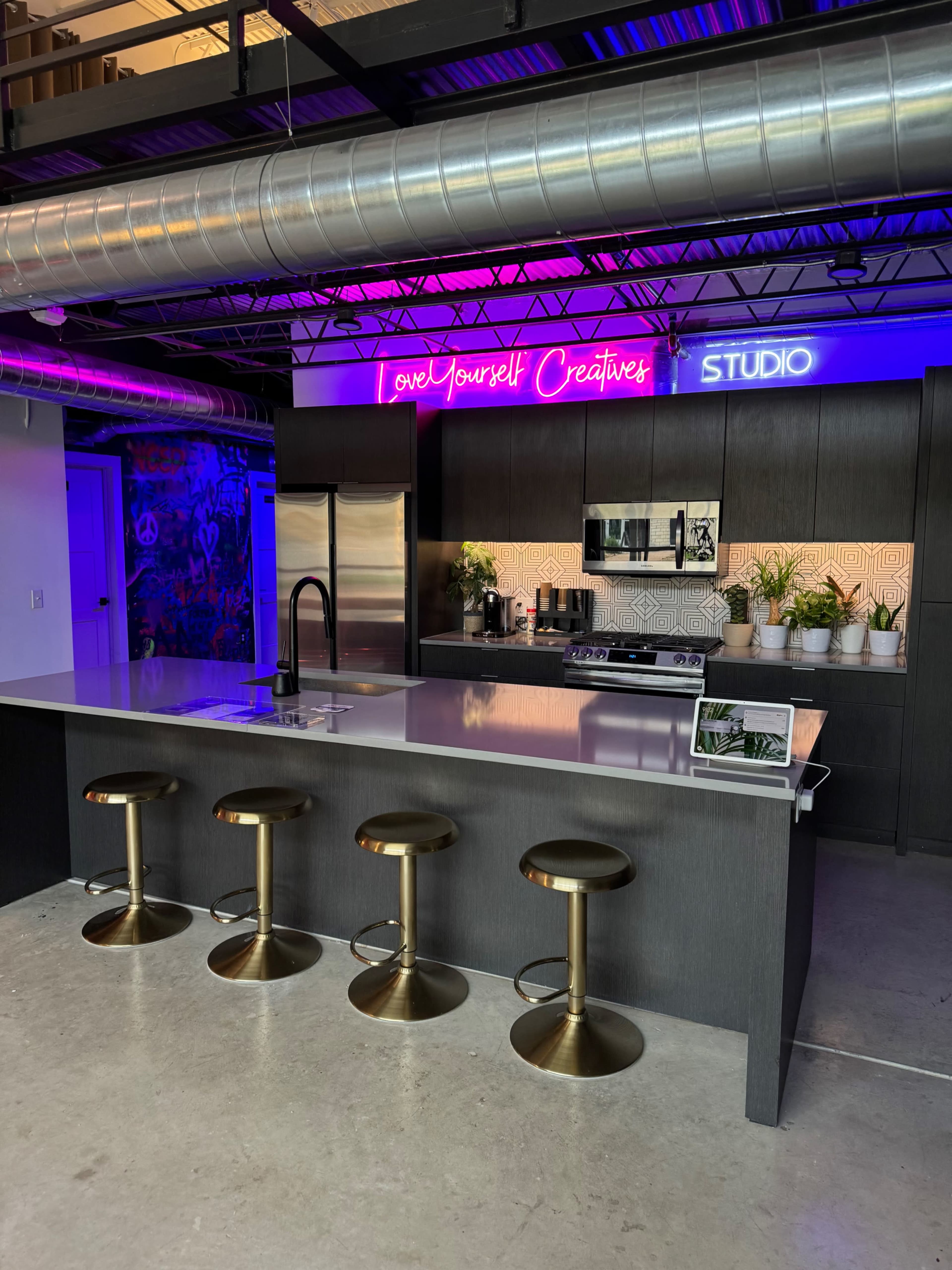 A modern kitchen features a large island with four brass stools, stainless steel appliances, and a neon sign that reads "Love Yourself Creative."
