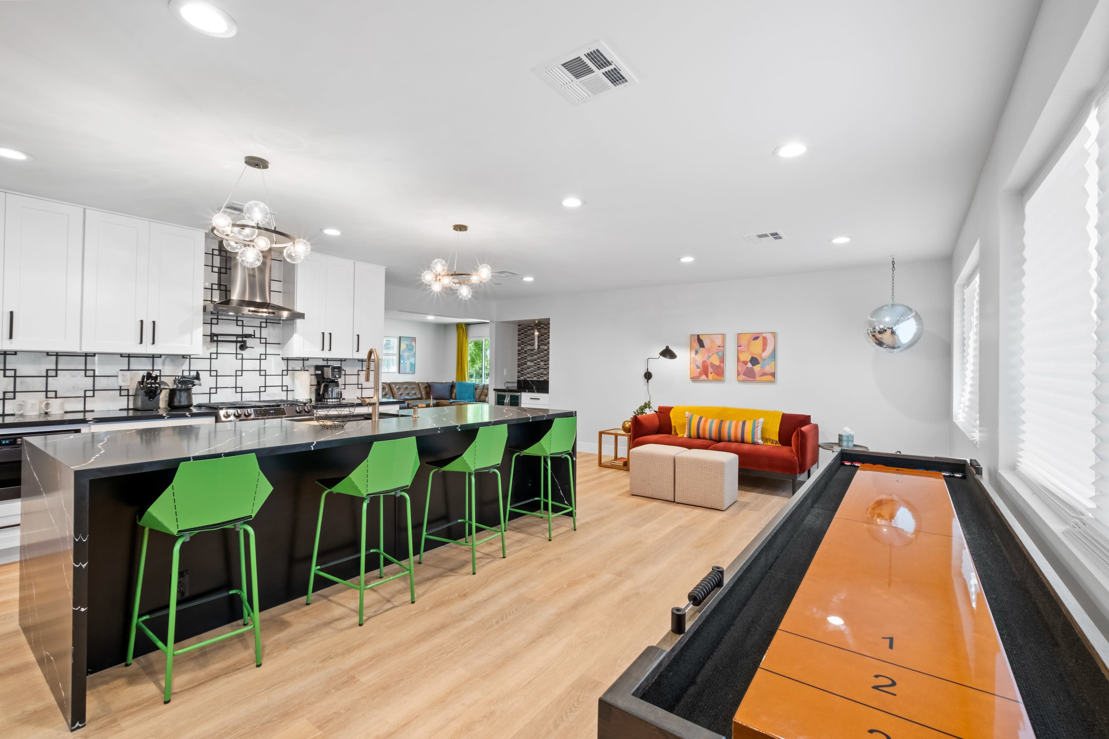 A modern kitchen with a large island, bright green bar stools, and a shuffleboard table in an open living space featuring a vibrant sofa and colorful artwork on the walls.