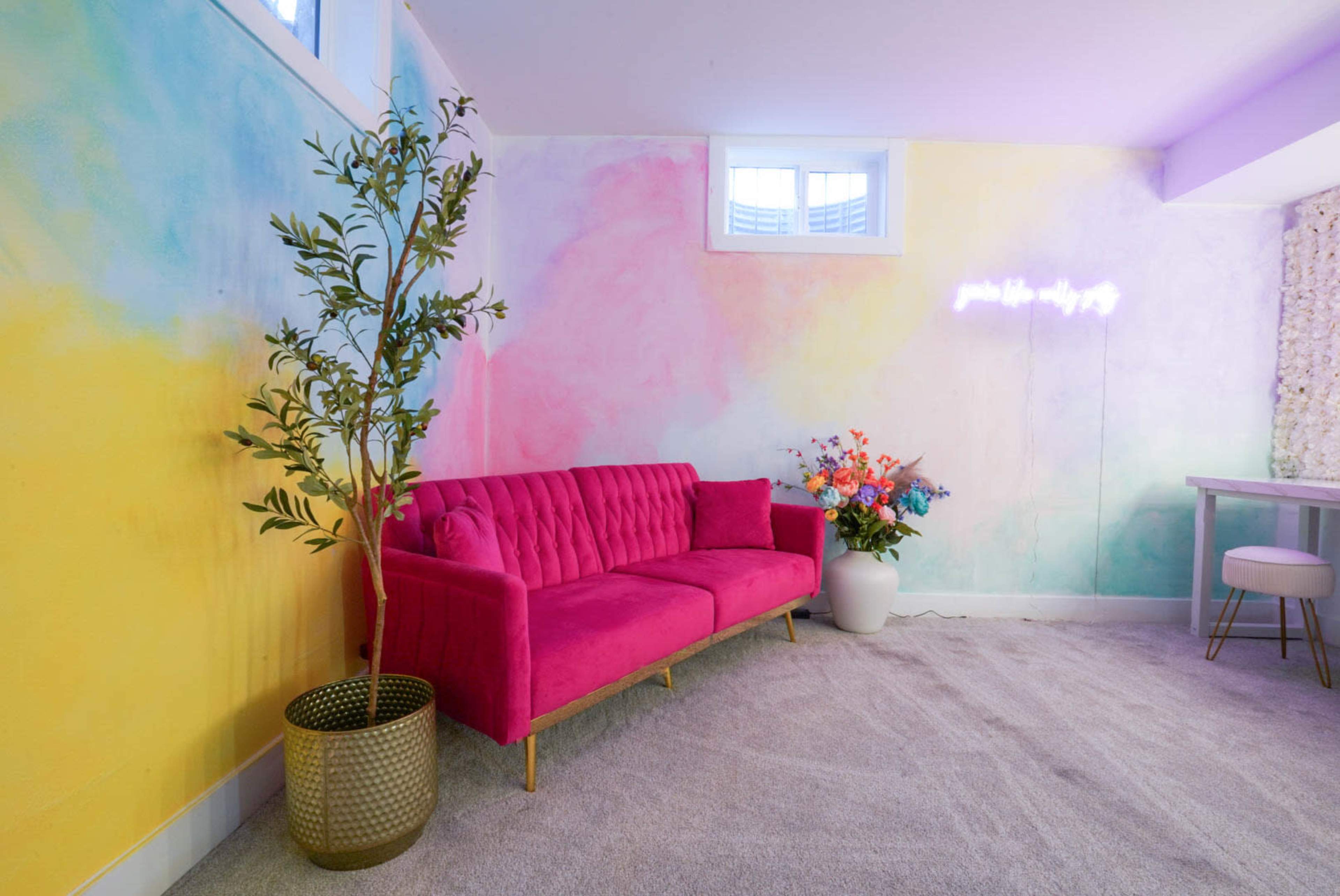 A bright, colorful room featuring a pink velvet sofa, a potted plant, and a decorative table against a pastel mural wall.