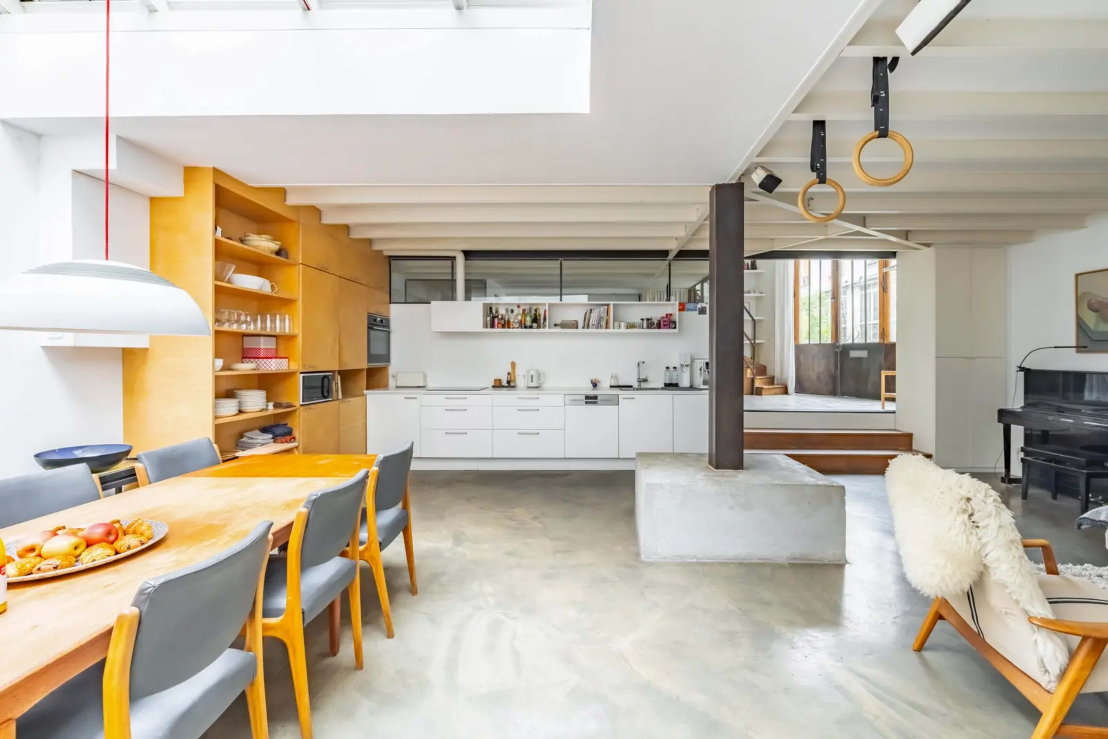 The scene depicts a modern kitchen and dining area featuring a large wooden dining table, white cabinetry, and minimalistic decor, with a visible staircase in the background leading to another level.