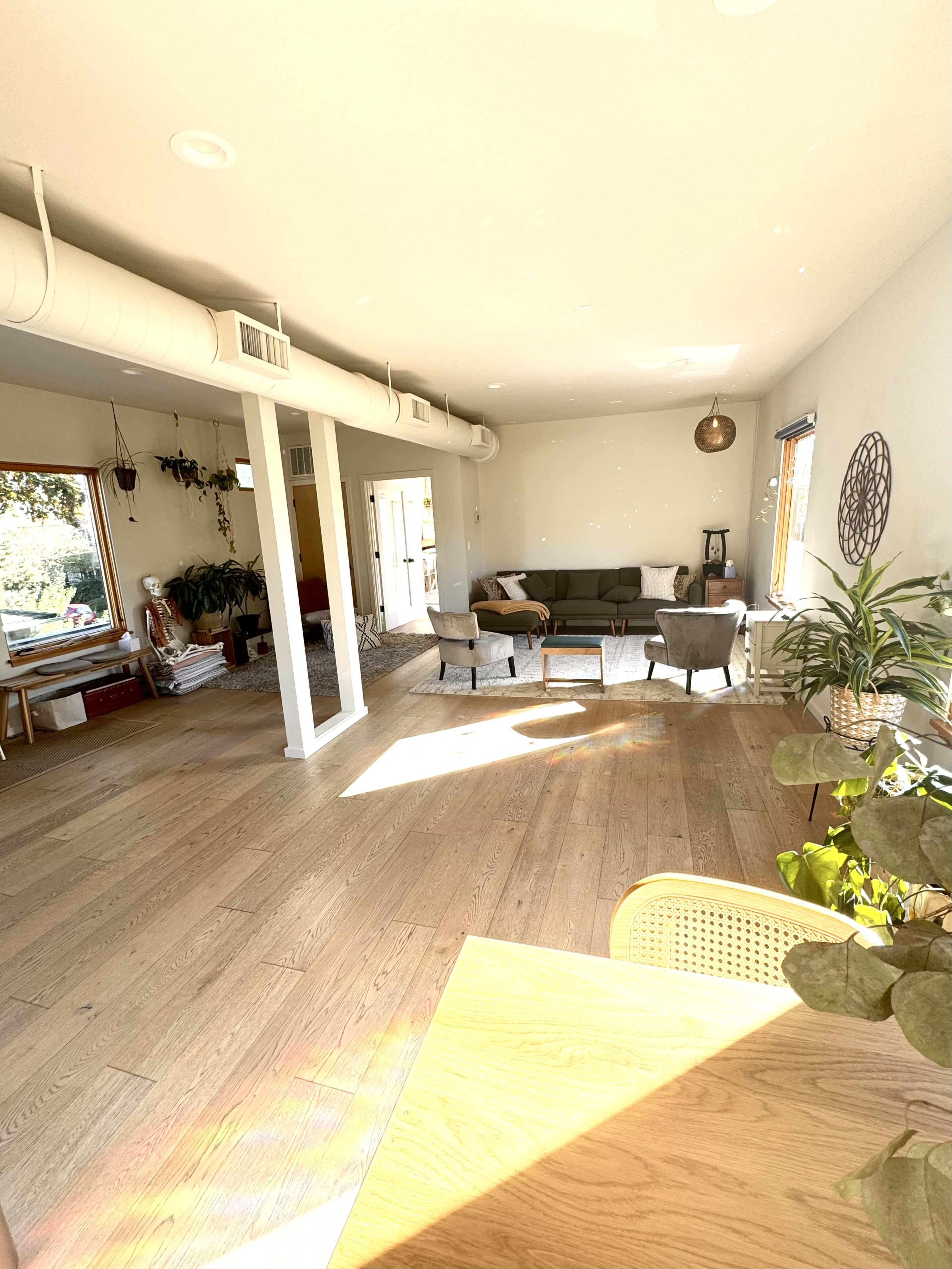 The image shows a spacious living area with wooden floors, a green sofa, and several chairs, illuminated by natural light from large windows.
