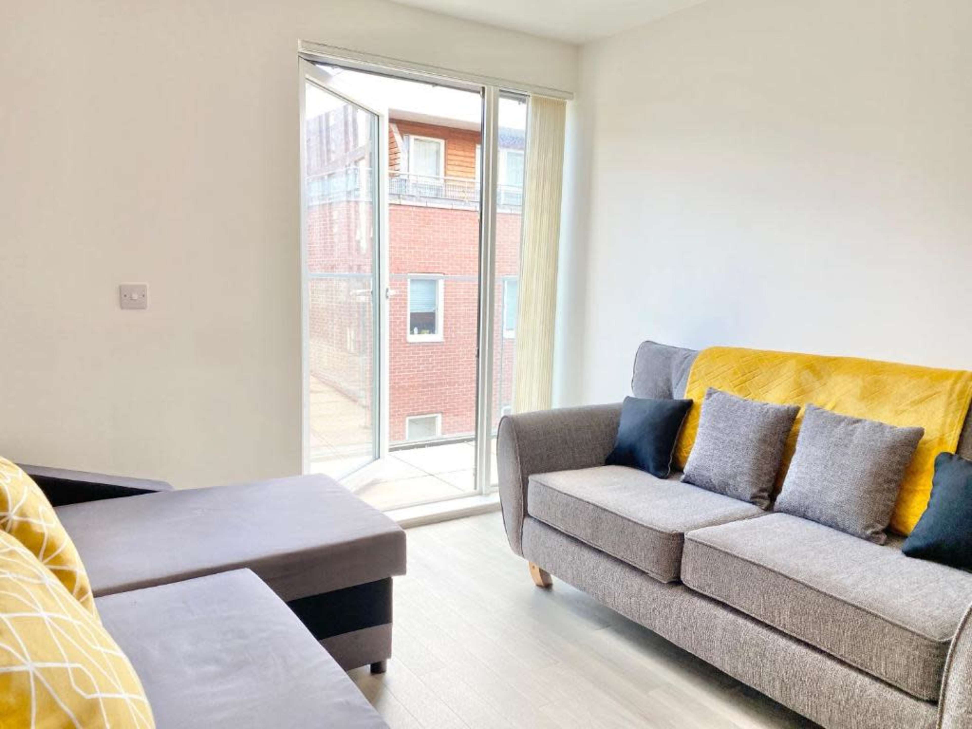 A bright, modern living room with a gray sofa, a yellow throw blanket, and large windows allowing natural light to enter.