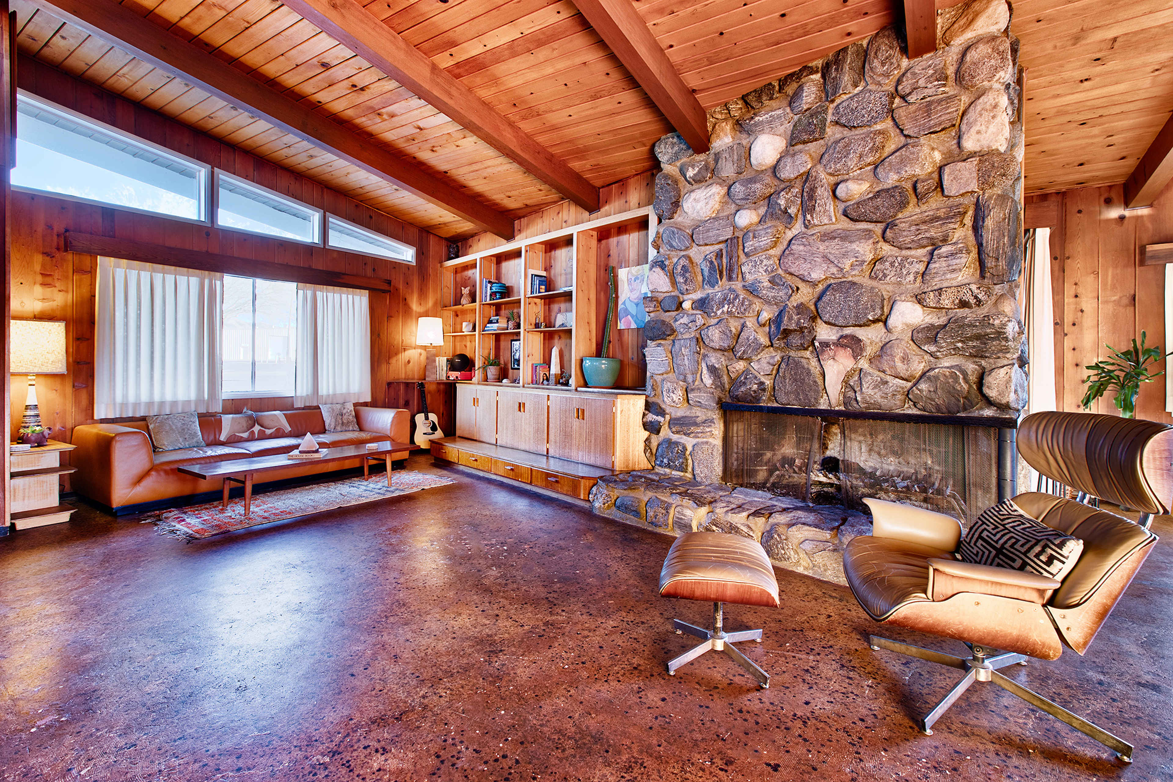 A spacious living room features a stone fireplace, wooden beams, and modern furniture arranged around a central coffee table.