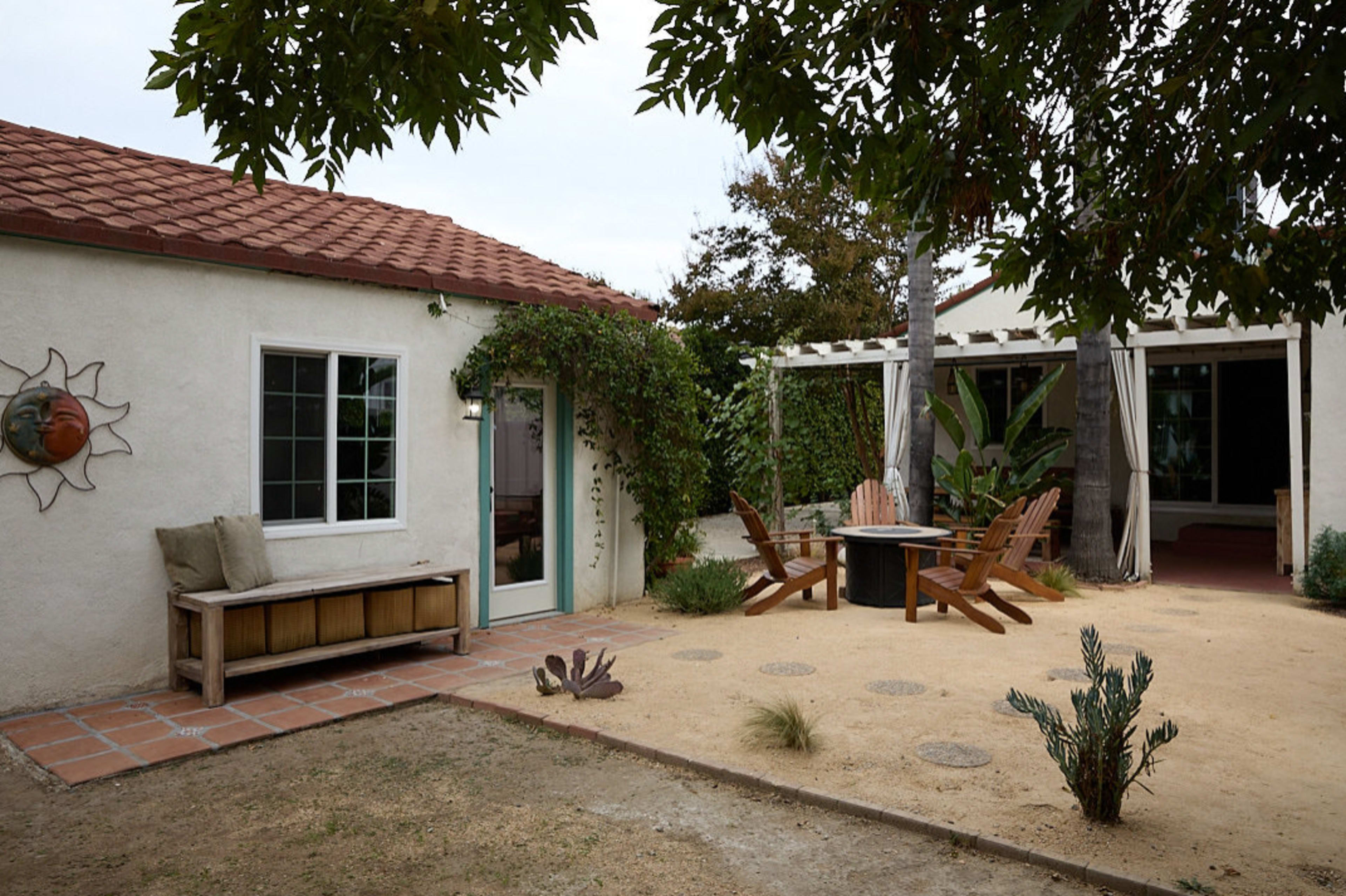 A small house with a red tiled roof features a wooden bench and a fire pit surrounded by seating in a landscaped yard.