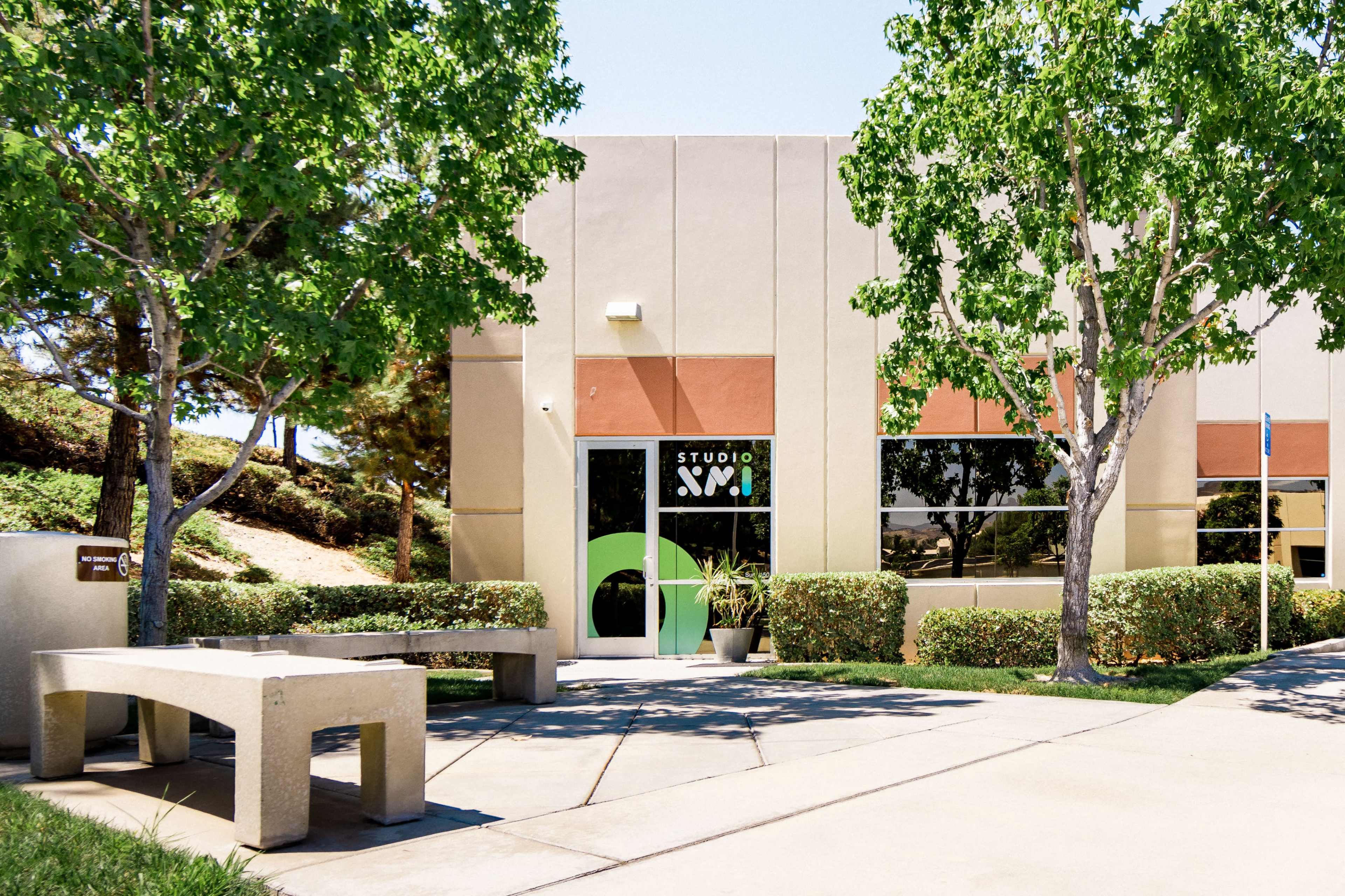 The image shows the exterior of a building labeled "STUDIO SVM," featuring large windows surrounded by green trees and neatly trimmed hedges.