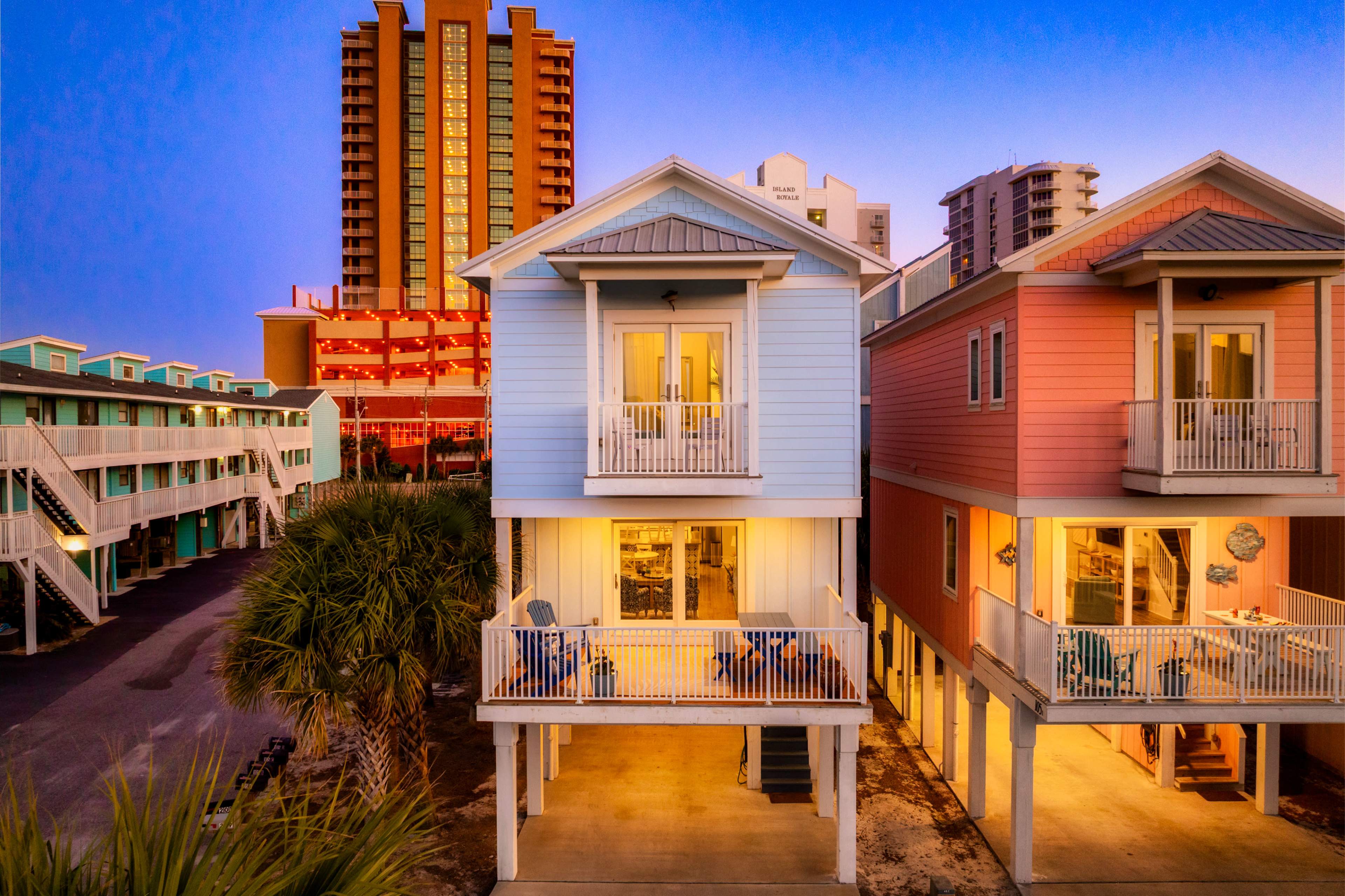 The image shows two brightly colored beach houses with balconies, surrounded by palm trees and taller buildings in the background during twilight.