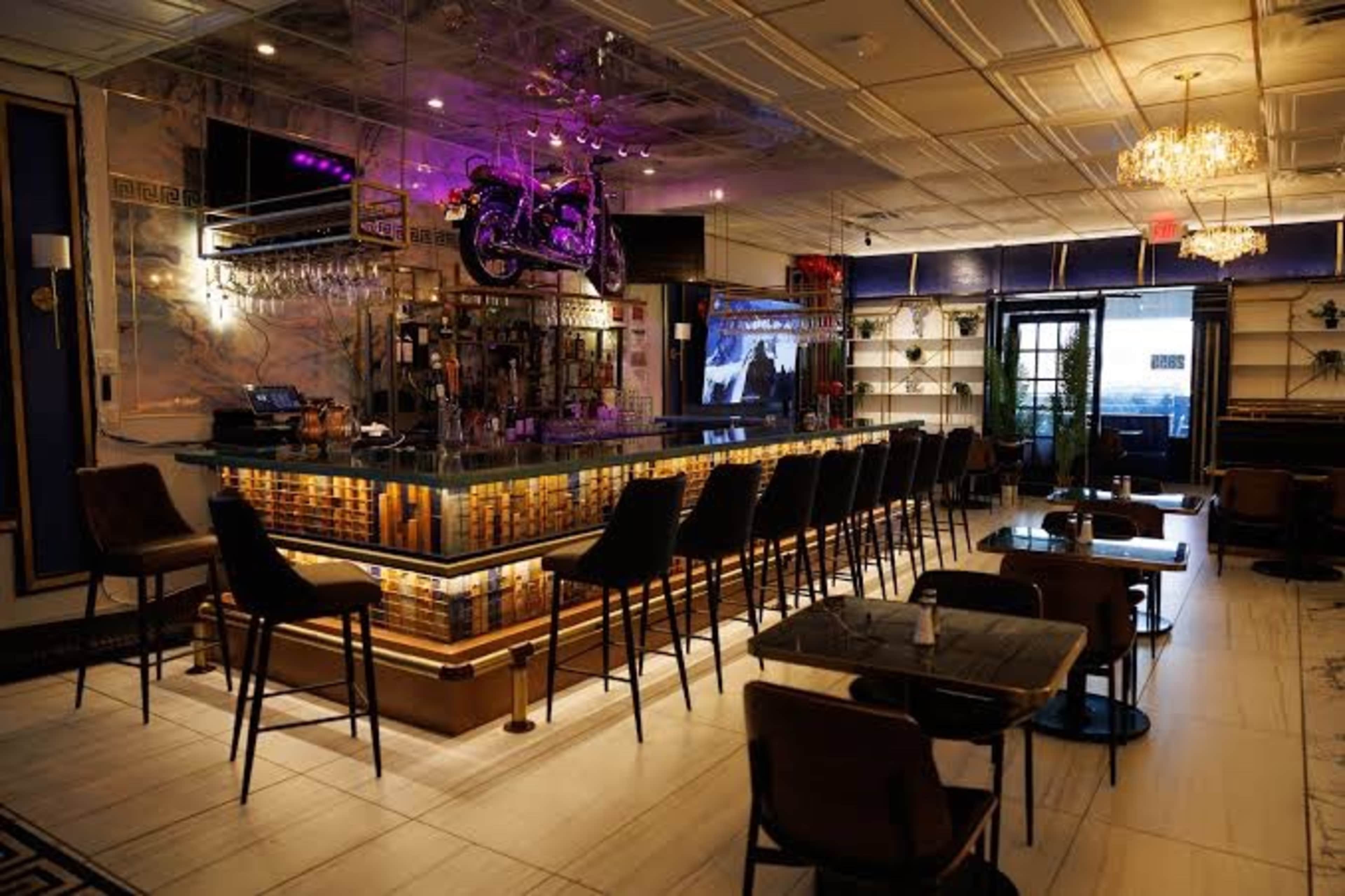The image shows a stylish bar with high stools, a well-lit counter featuring decorative elements, and a motorcycle suspended above it, set in a modern interior space.