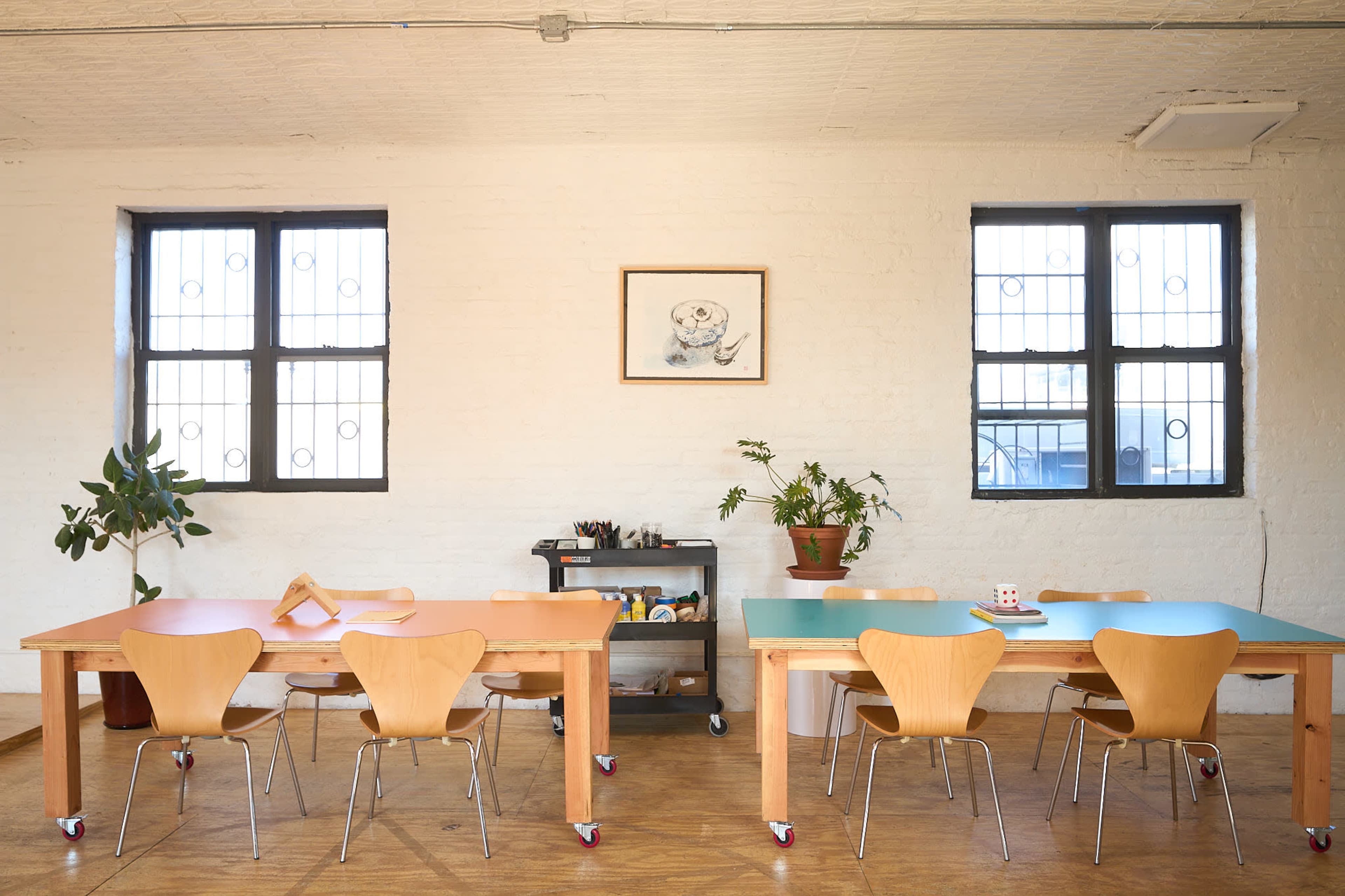 The image shows a well-lit room with two large wooden tables surrounded by wooden chairs, a potted plant, and artwork on the wall.