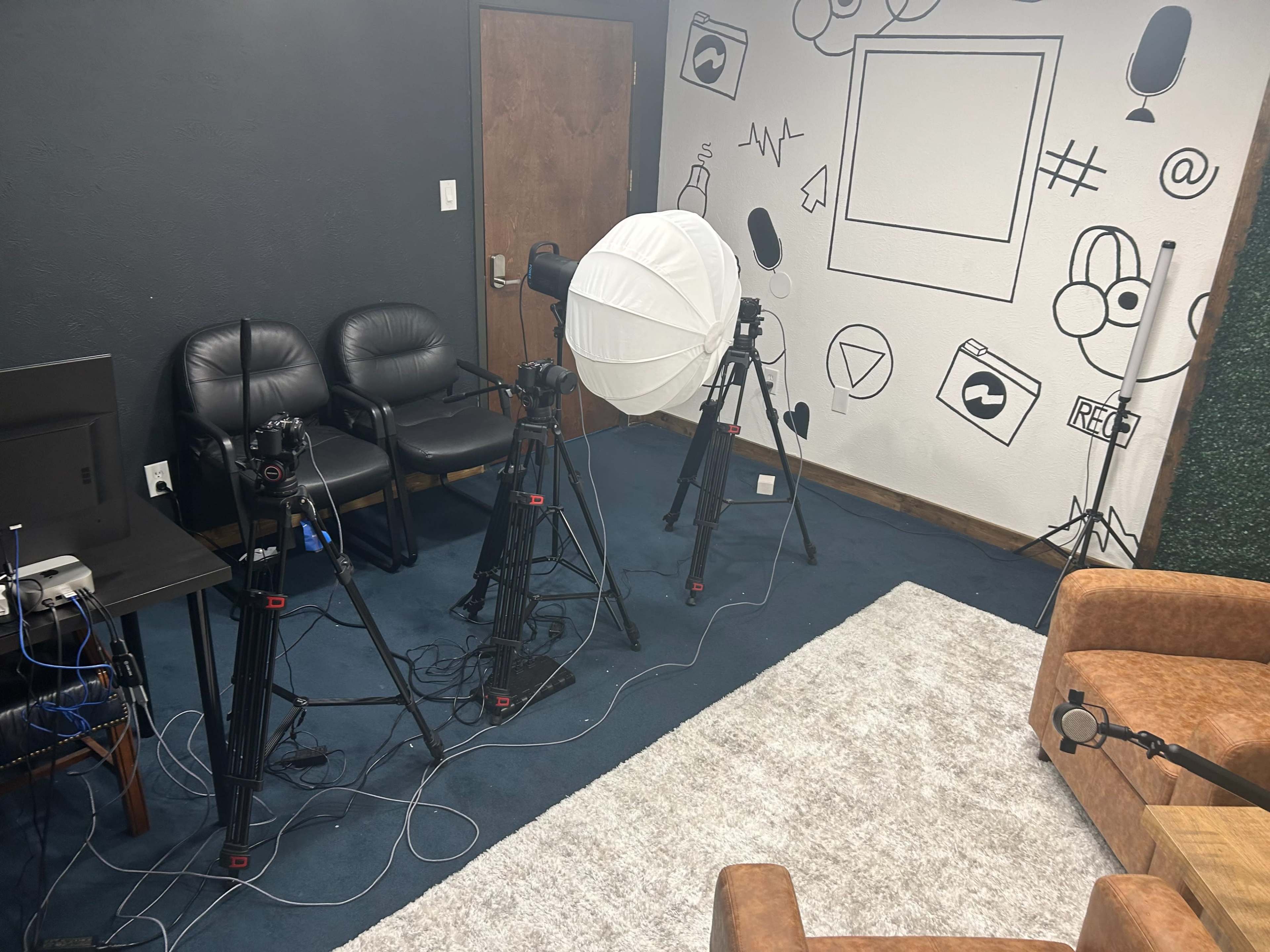 The image shows a small recording studio with three camera tripods positioned in front of a blank wall adorned with various graphic designs, along with two seating areas and a monitor in the corner.