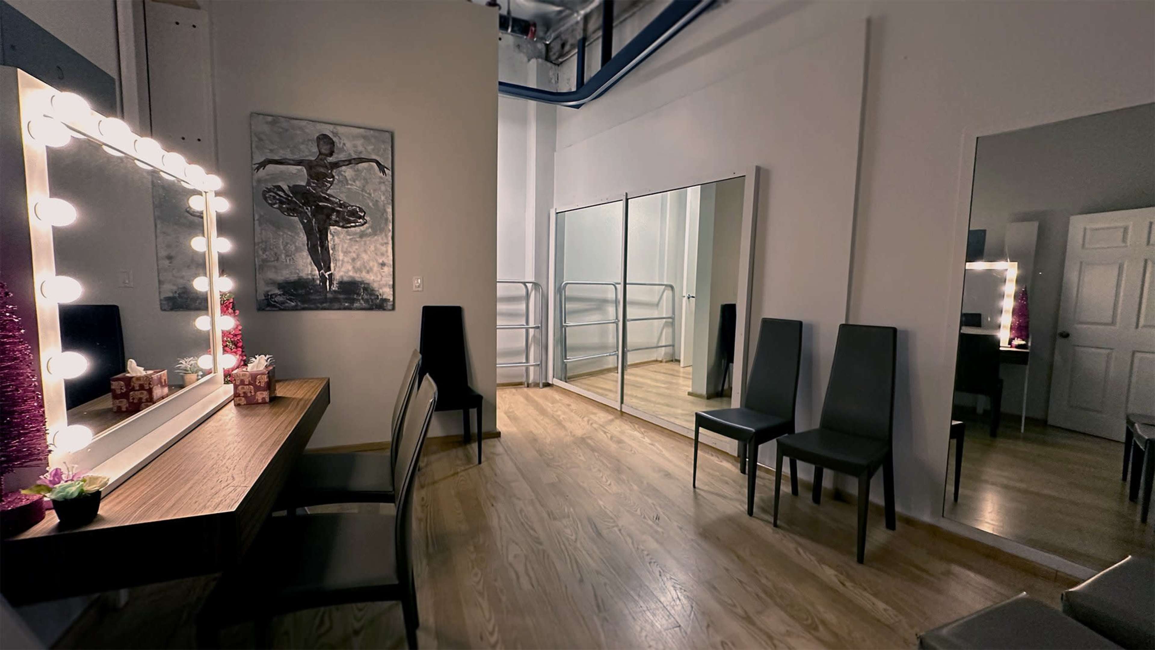 A spacious dressing room with a lighted vanity mirror, several chairs, and a large mirrored wall for practice.