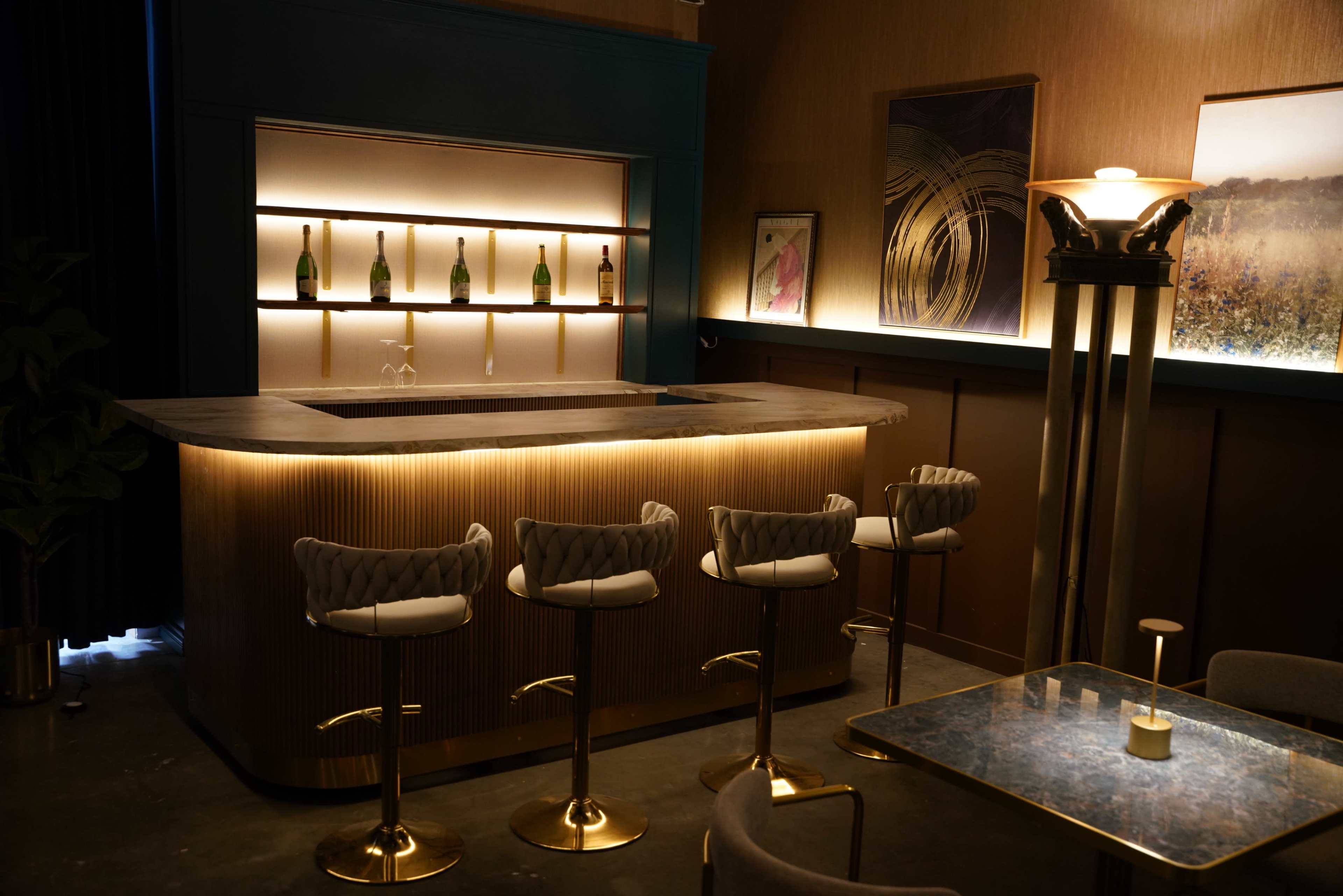 The image shows a modern bar area with a sleek counter, stylish stools, and illuminated shelves holding several bottles.