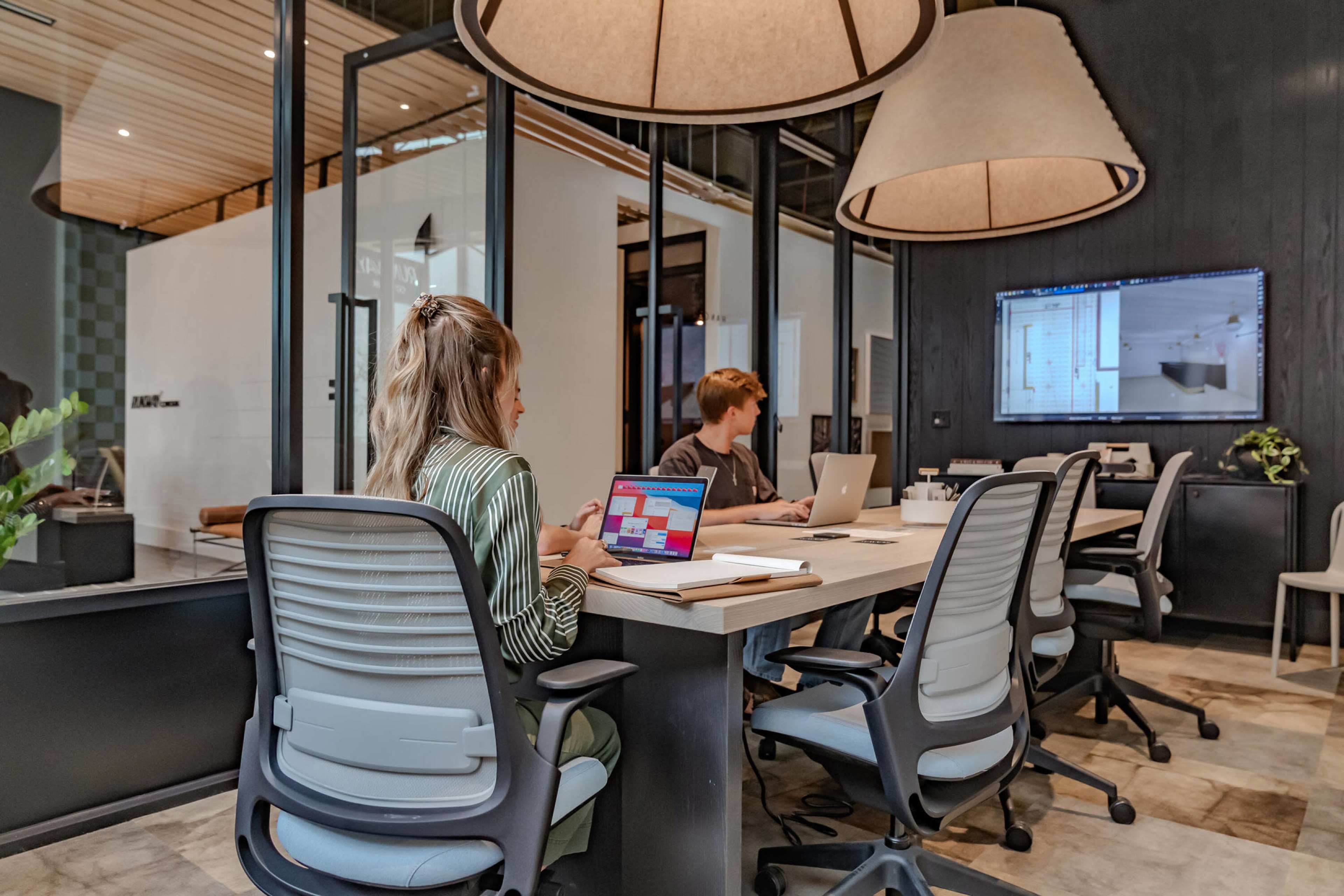 Two people are working at a modern office table equipped with laptops in a well-lit conference room.
