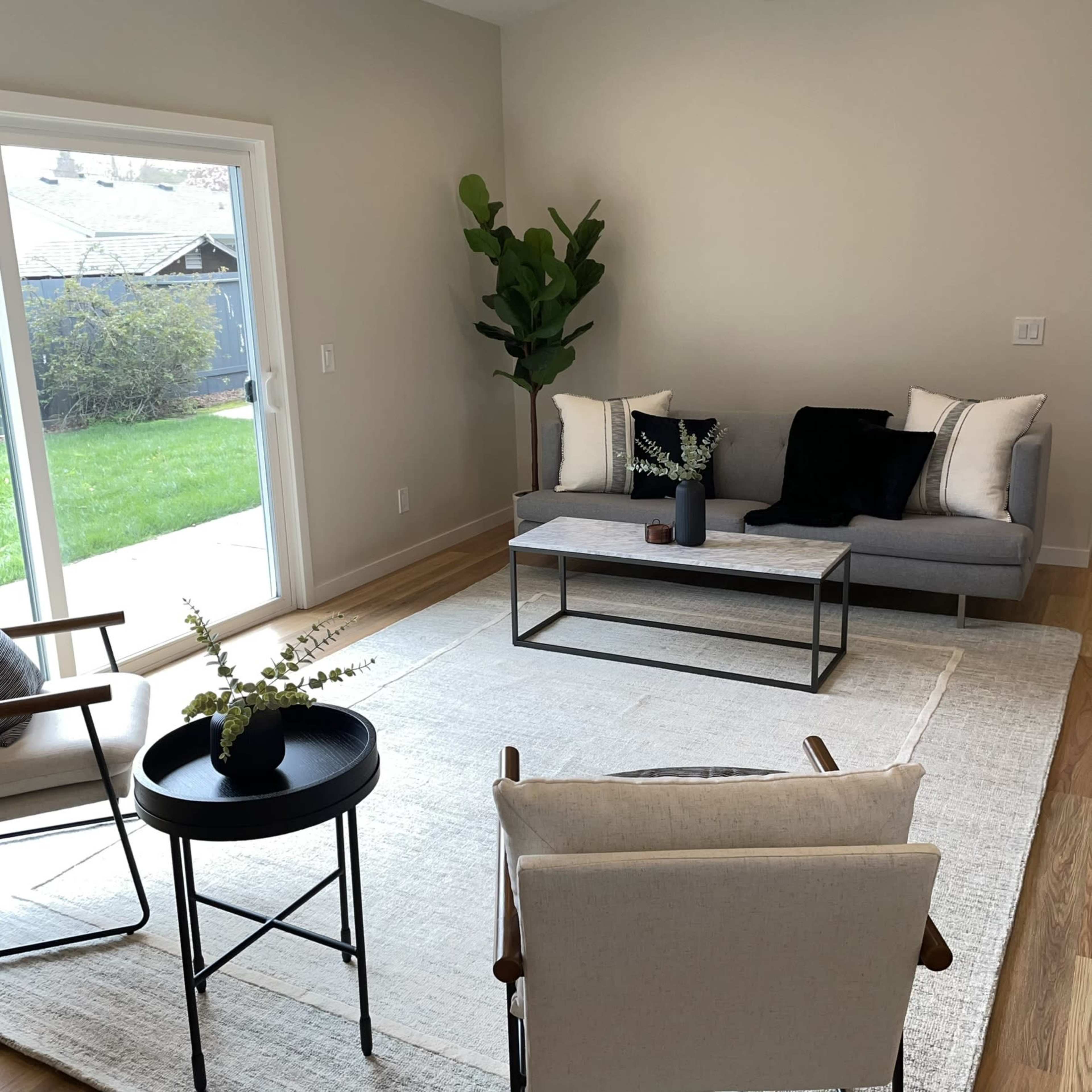 The scene shows a modern living room with a gray sofa, a glass coffee table, two armchairs, and a sliding glass door leading to a grassy outdoor space.