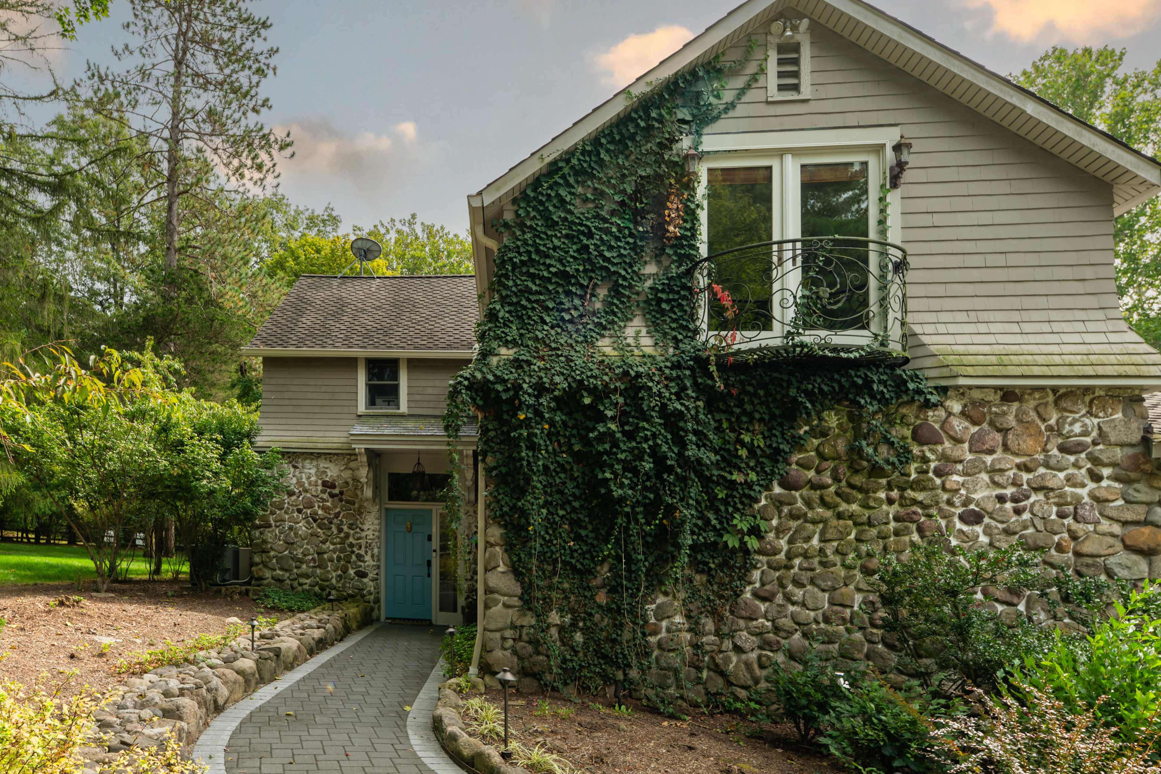 The image shows a two-story house with stone and wooden siding, featuring a blue front door and a balcony covered in green ivy.