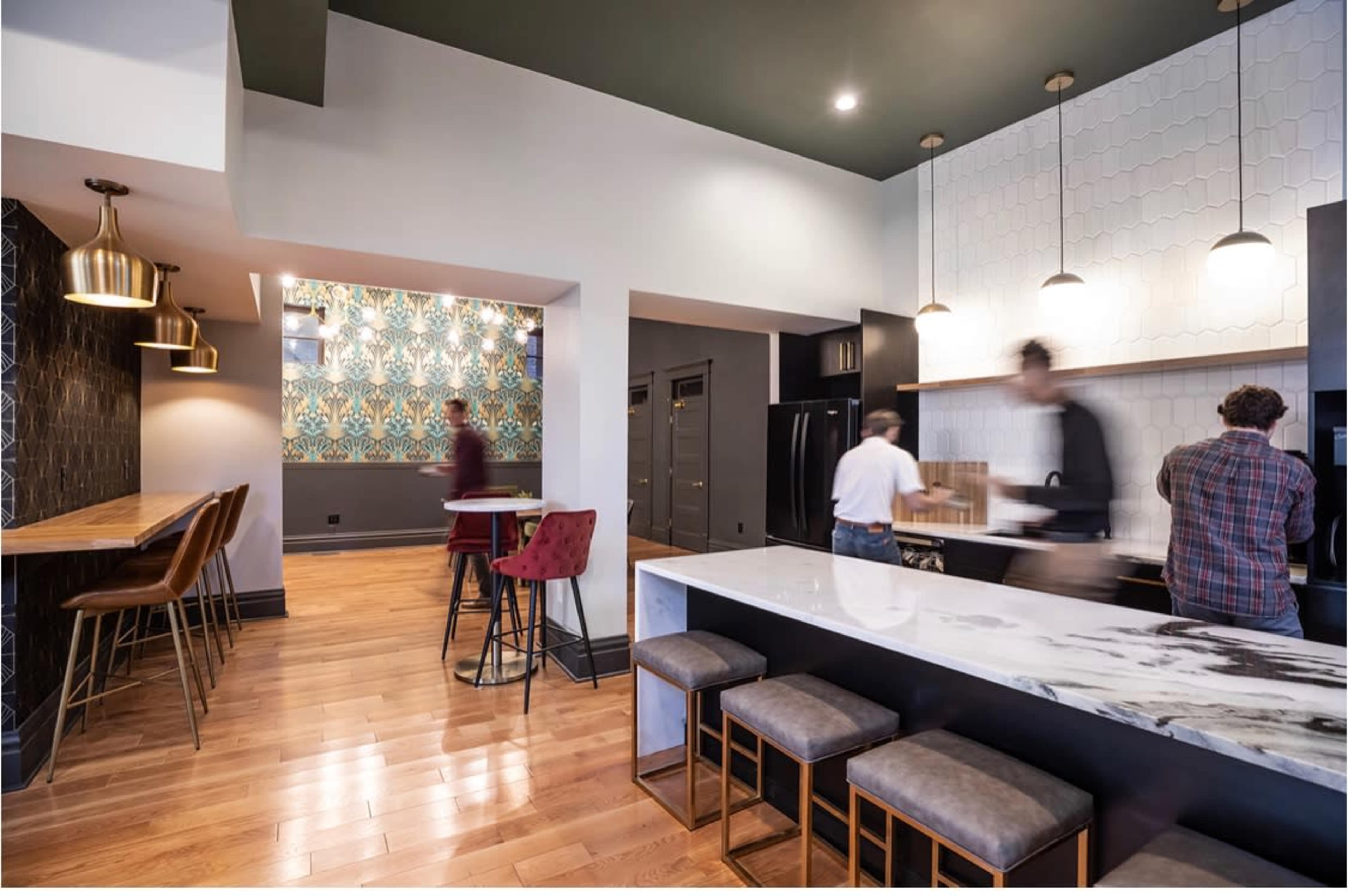 A modern kitchen with a large marble island, wooden flooring, and a patterned accent wall, while several people are engaged in activities in the space.