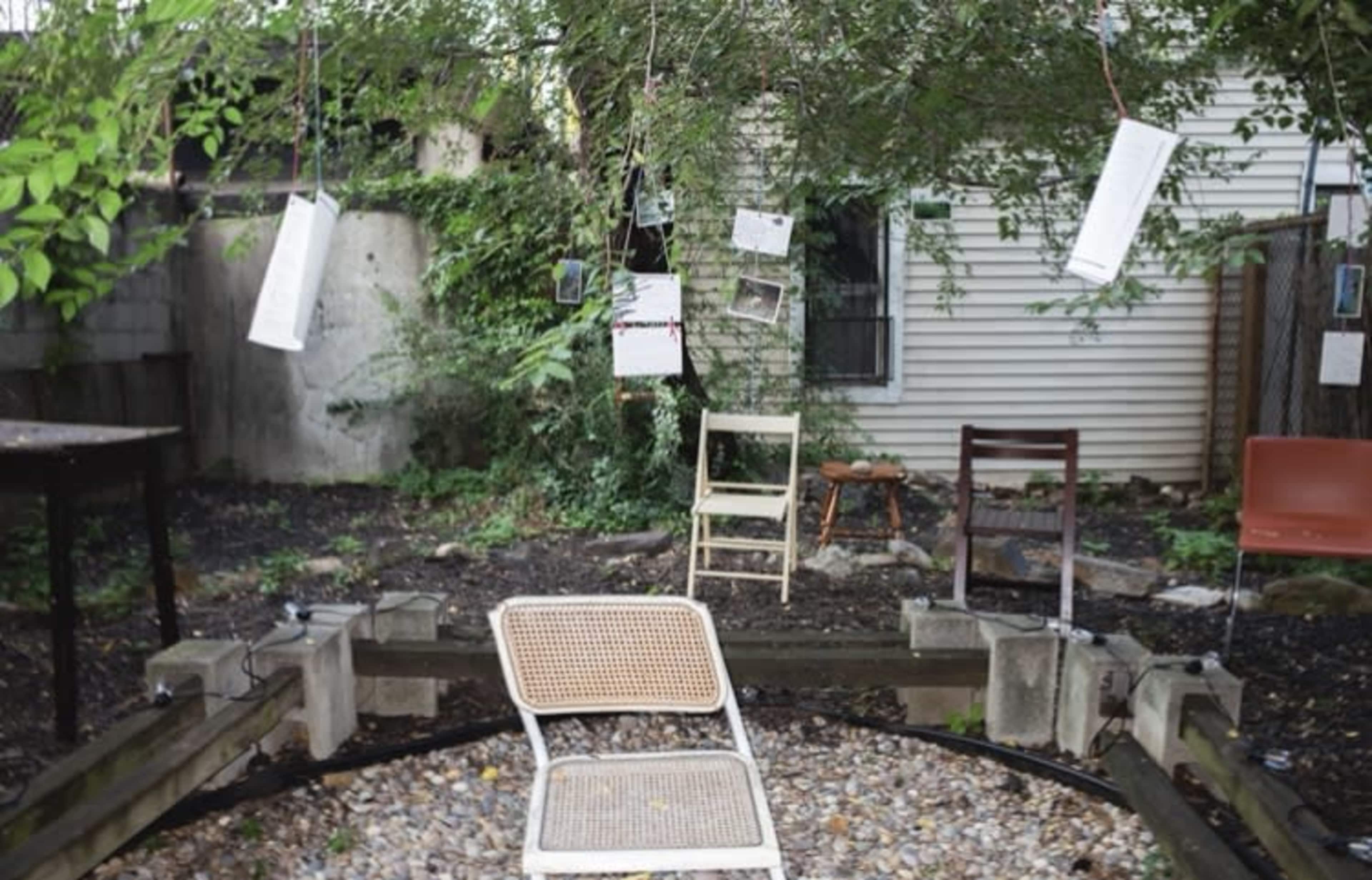The image shows a garden area with several chairs arranged around a circular gravel patch, while papers hang from tree branches above.