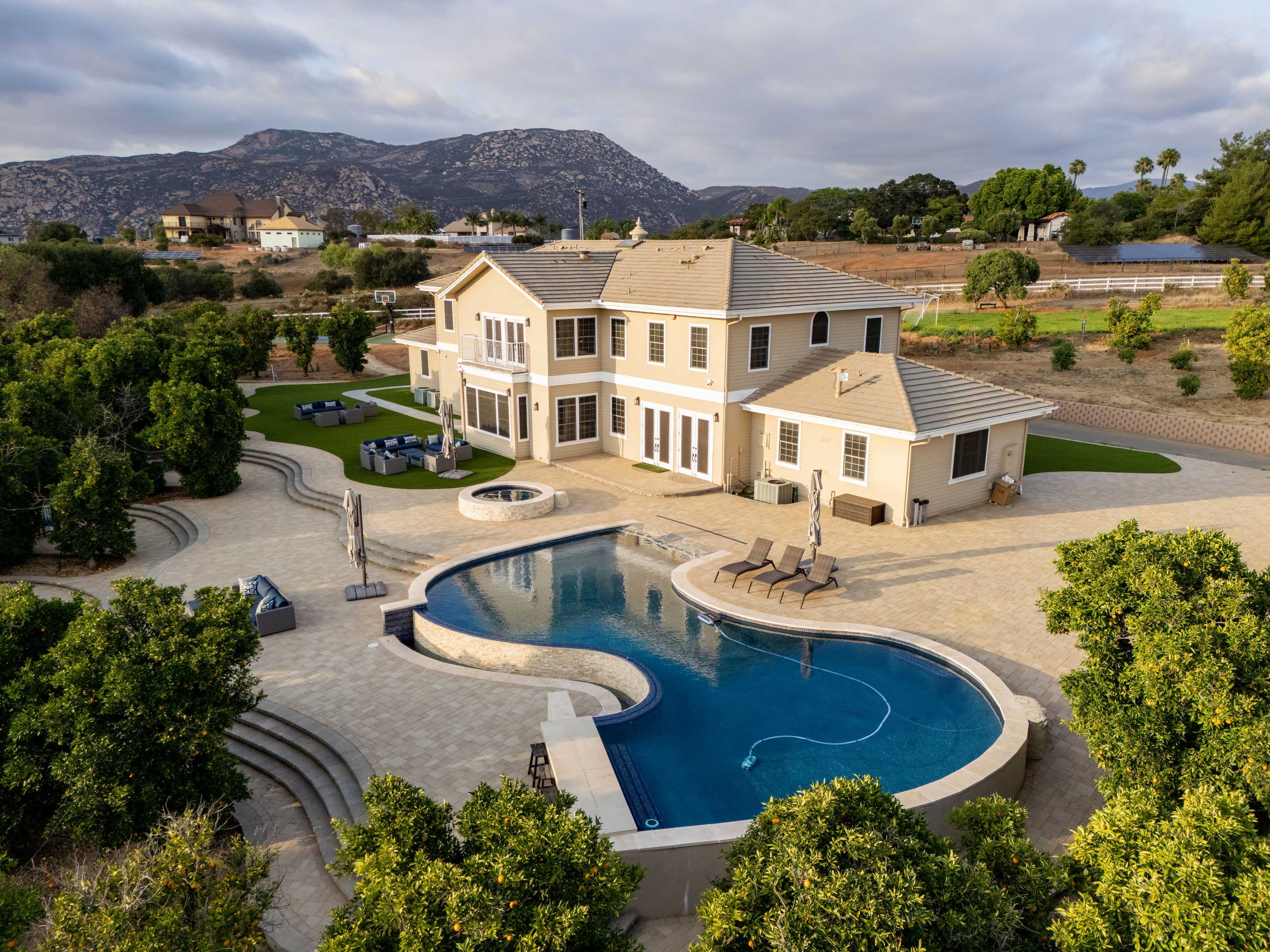 The image shows a large two-story house with an expansive backyard featuring a swimming pool, lounge chairs, and surrounding landscaping, set against a backdrop of hills.