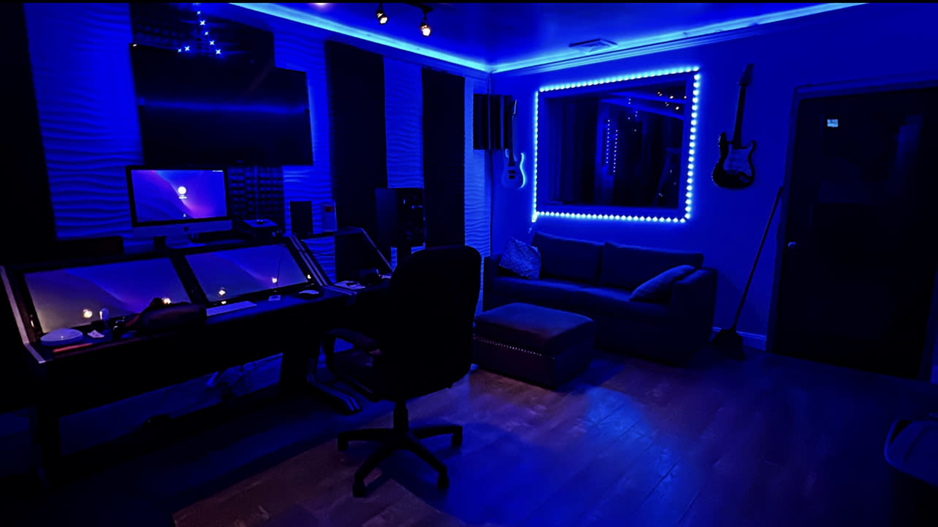 A music studio with blue LED lighting, featuring a desk with multiple screens, a chair, a sofa, and a guitar mounted on the wall.