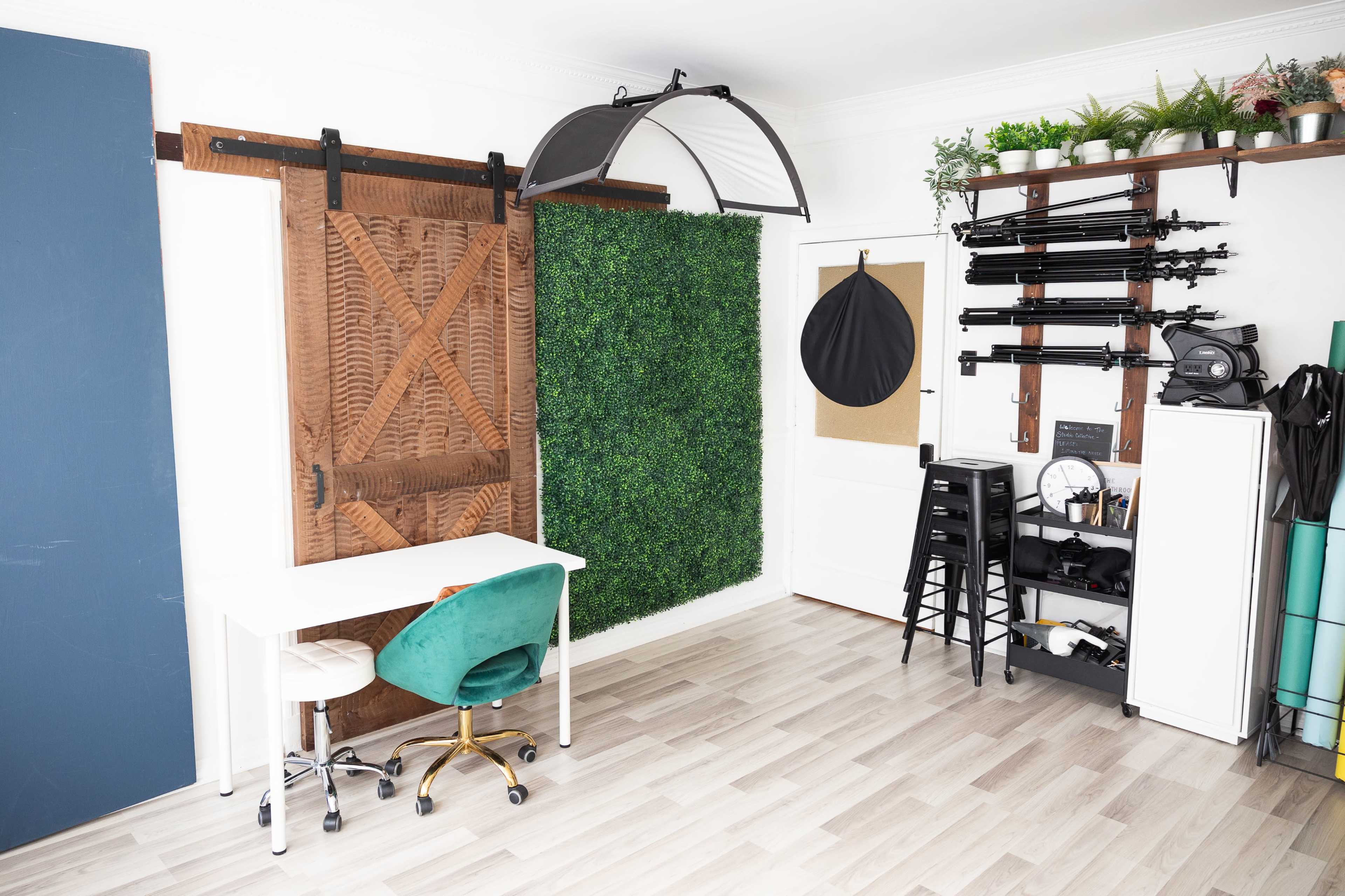 A modern workspace featuring a white desk with a green chair, a large wooden sliding door, a wall covered in artificial greenery, and various photography equipment mounted on the opposite wall.