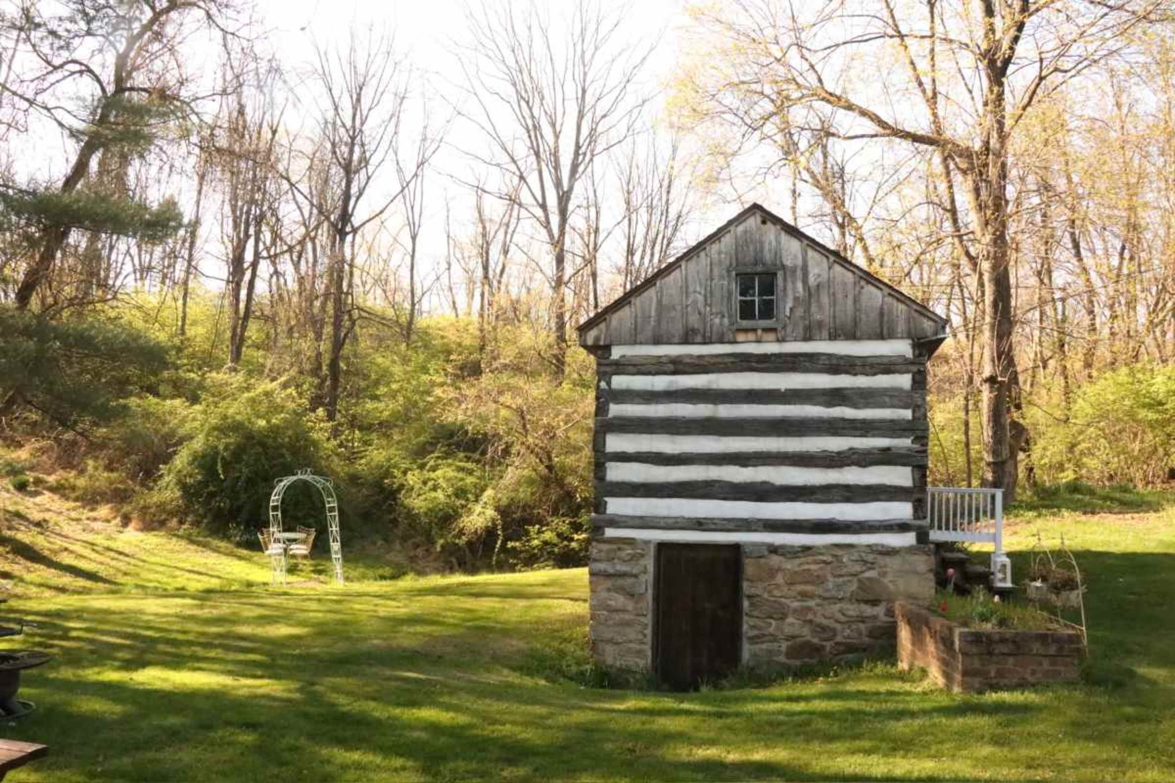 The image features a rustic, two-story log cabin surrounded by green grass and trees, with a bench swing nearby.