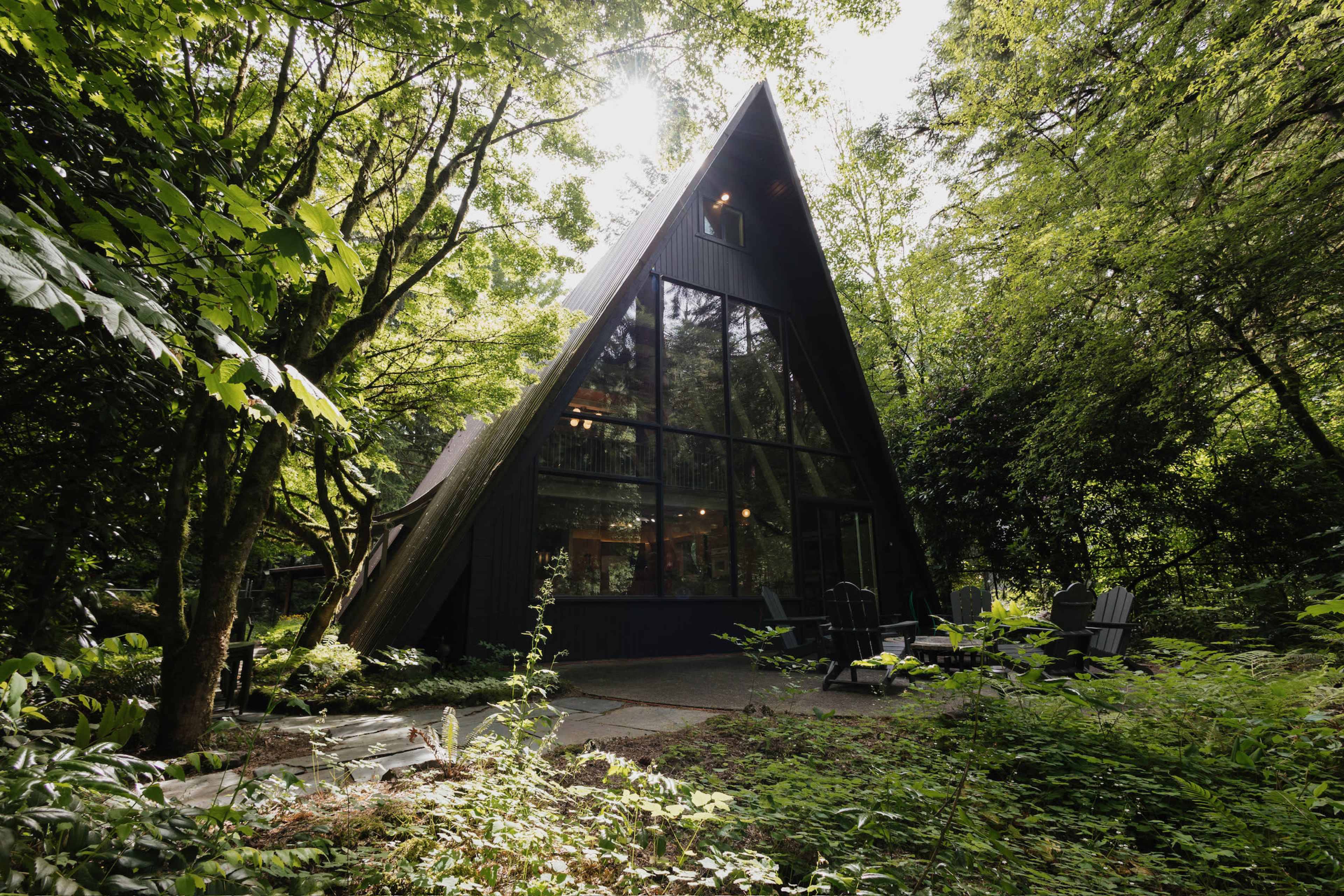 The image shows a modern A-frame house surrounded by tall trees and lush greenery.