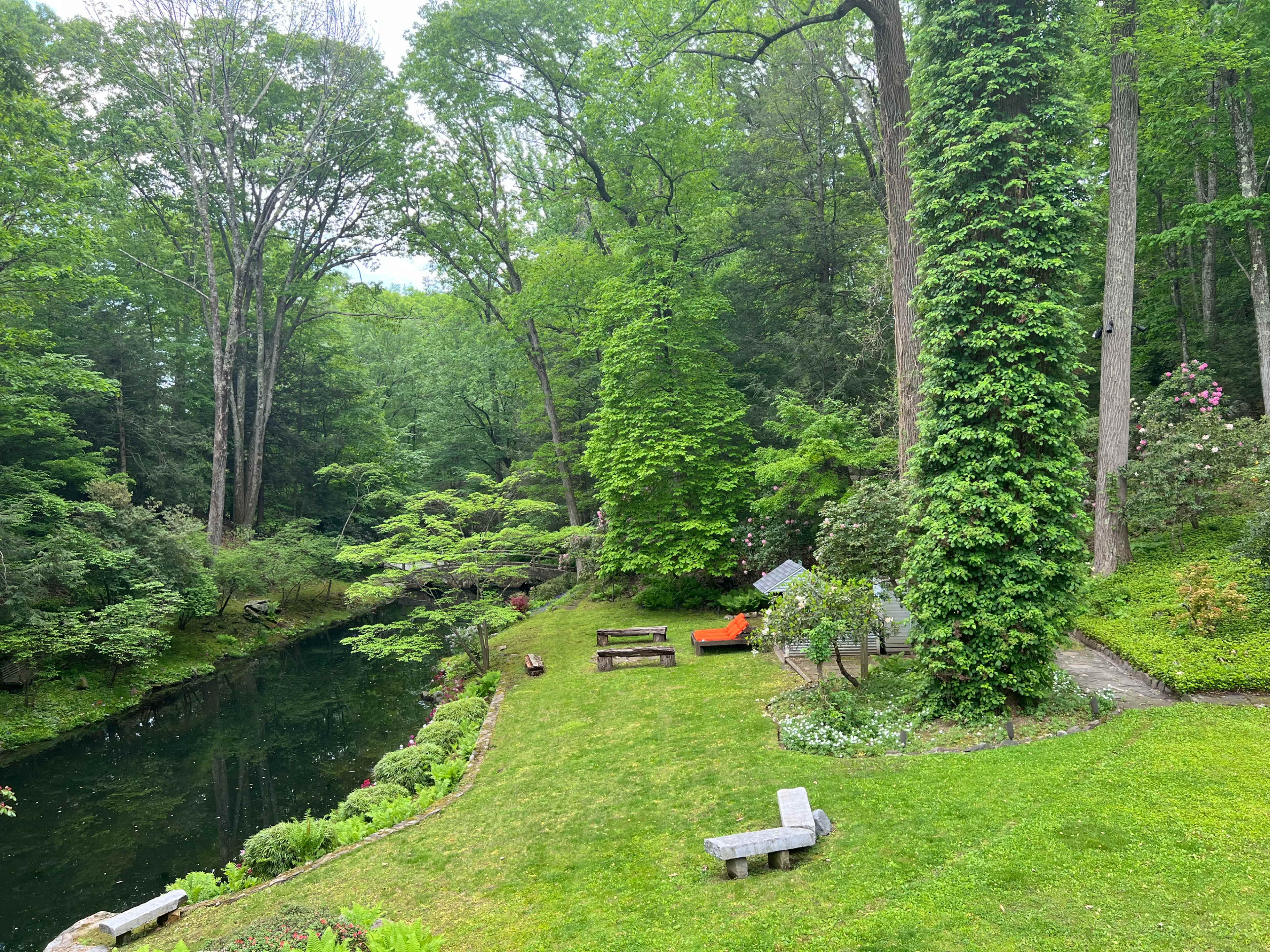 A lush green landscape with a pond, surrounded by trees, and a seating area with benches and an orange chair.