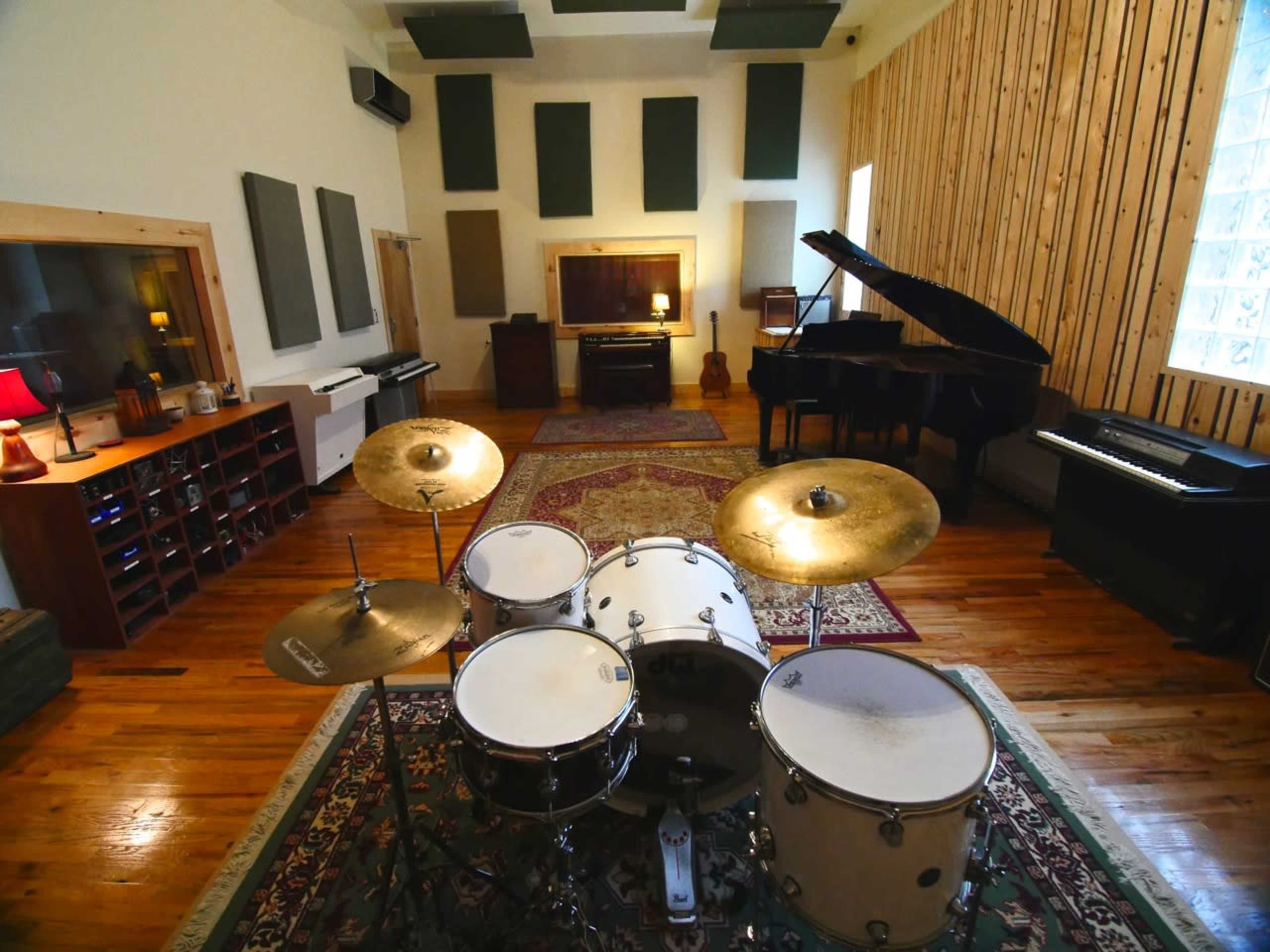 A spacious music studio featuring a drum set, a grand piano, and two upright pianos arranged against wooden walls and acoustic panels.