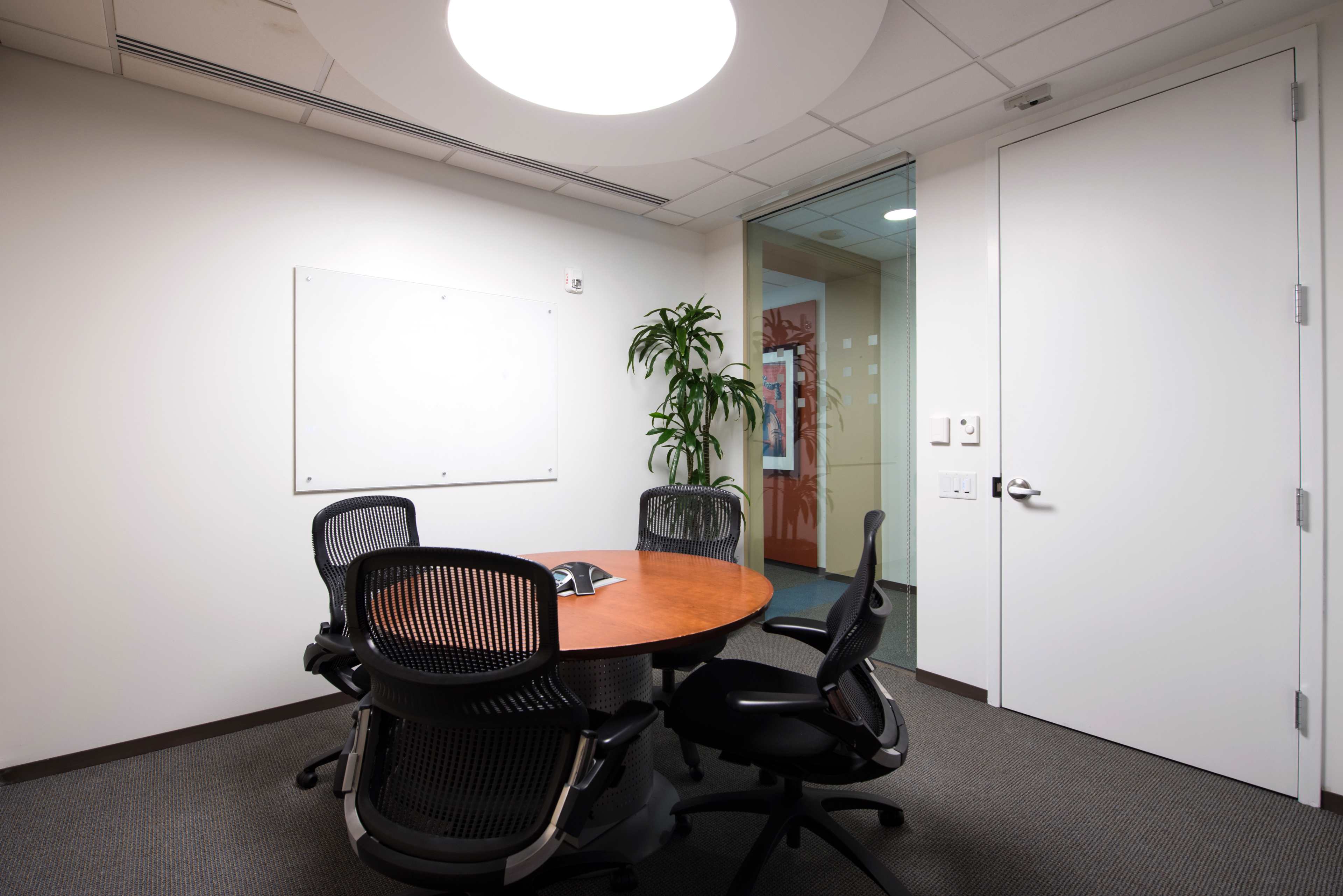 A small conference room features a round wooden table surrounded by four black mesh chairs, with a whiteboard on one wall and a glass wall leading to another room.