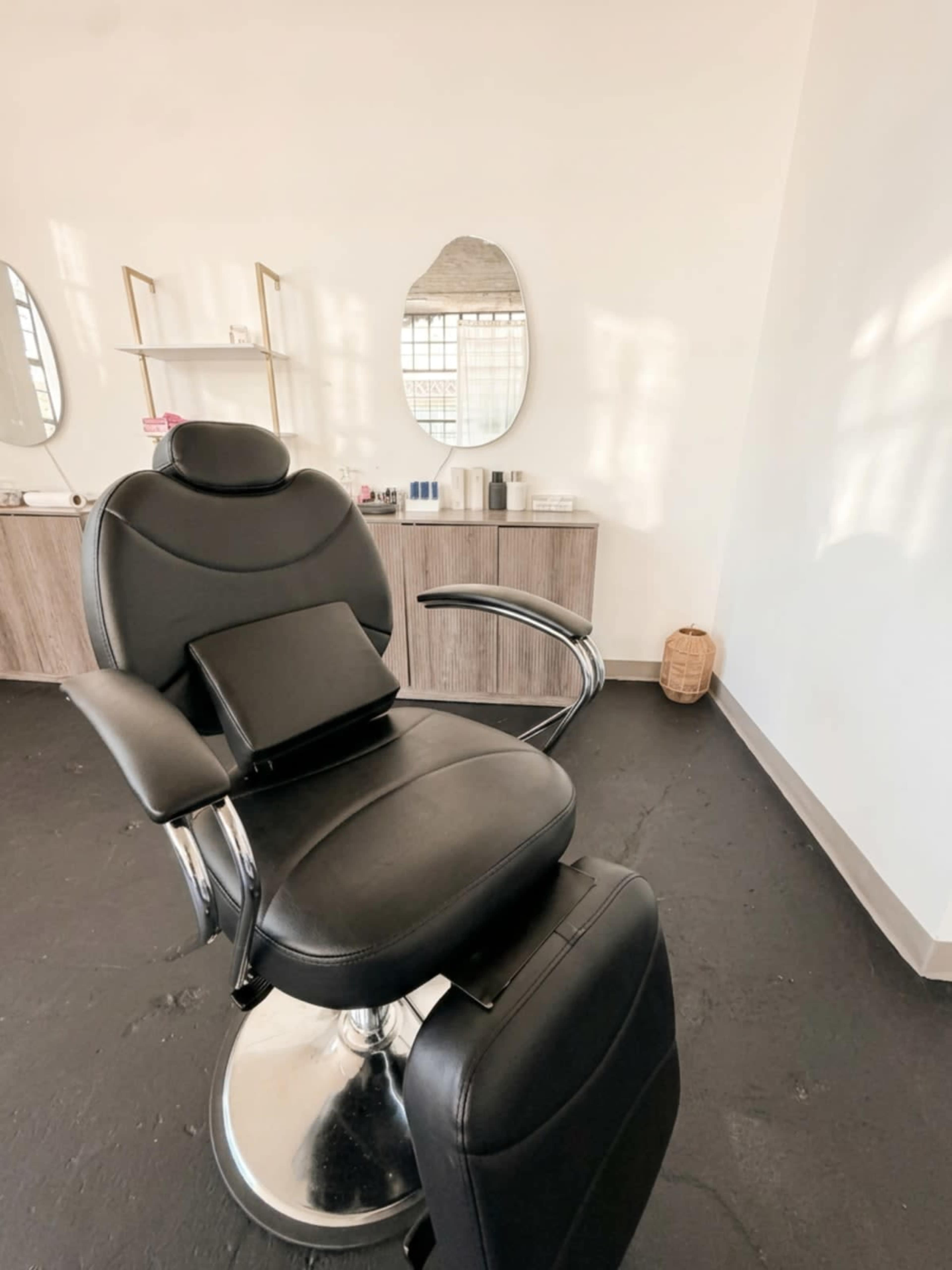 The image features a black salon chair in a minimalist beauty room with a mirror and a wooden storage unit in the background.