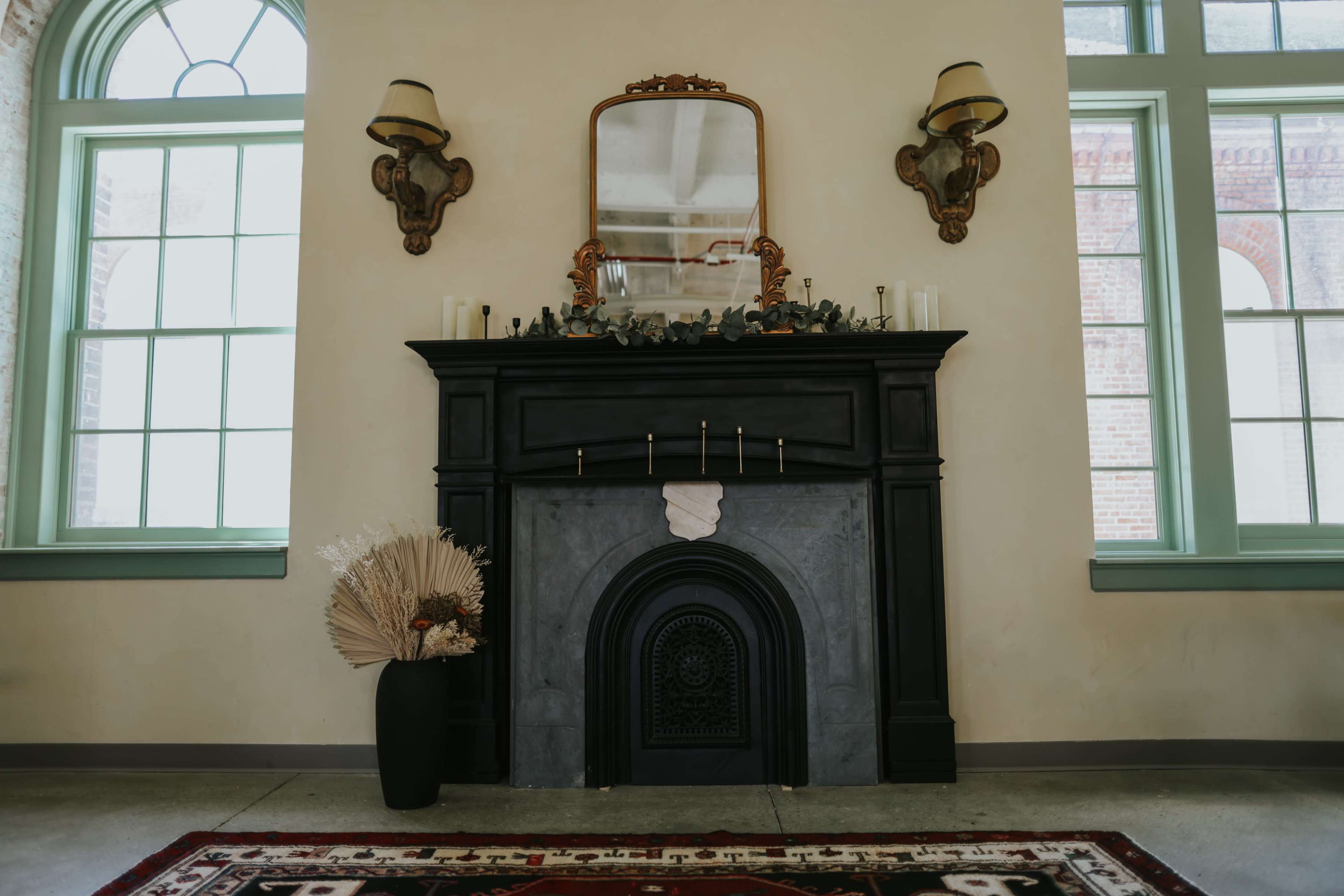 The image features a black fireplace with a mirror above it, flanked by wall sconces, against a light-colored wall, with a decorative vase and dried plants beside it.