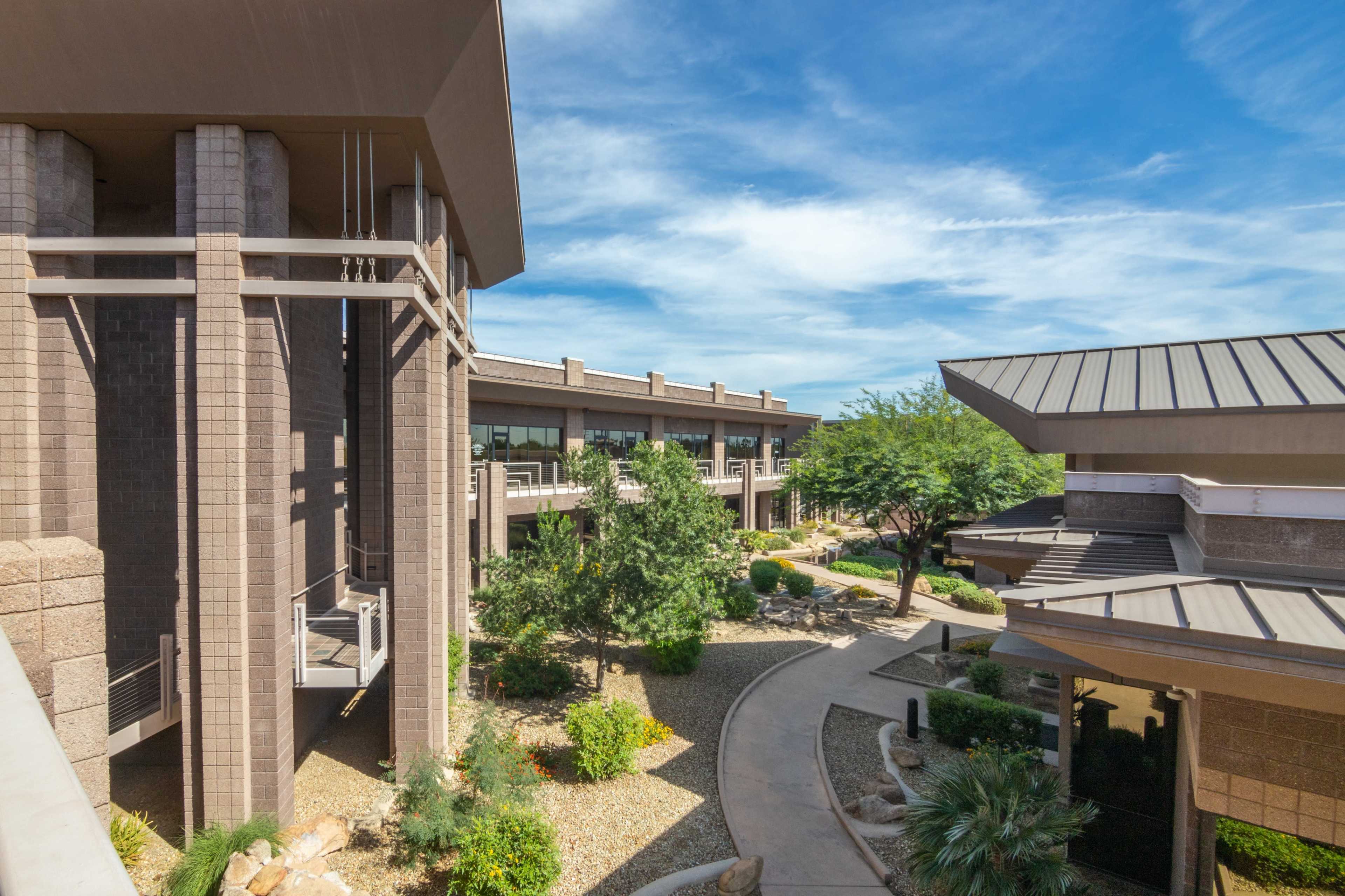The image shows a modern architectural complex with multiple buildings surrounded by landscaped pathways and greenery under a clear blue sky.