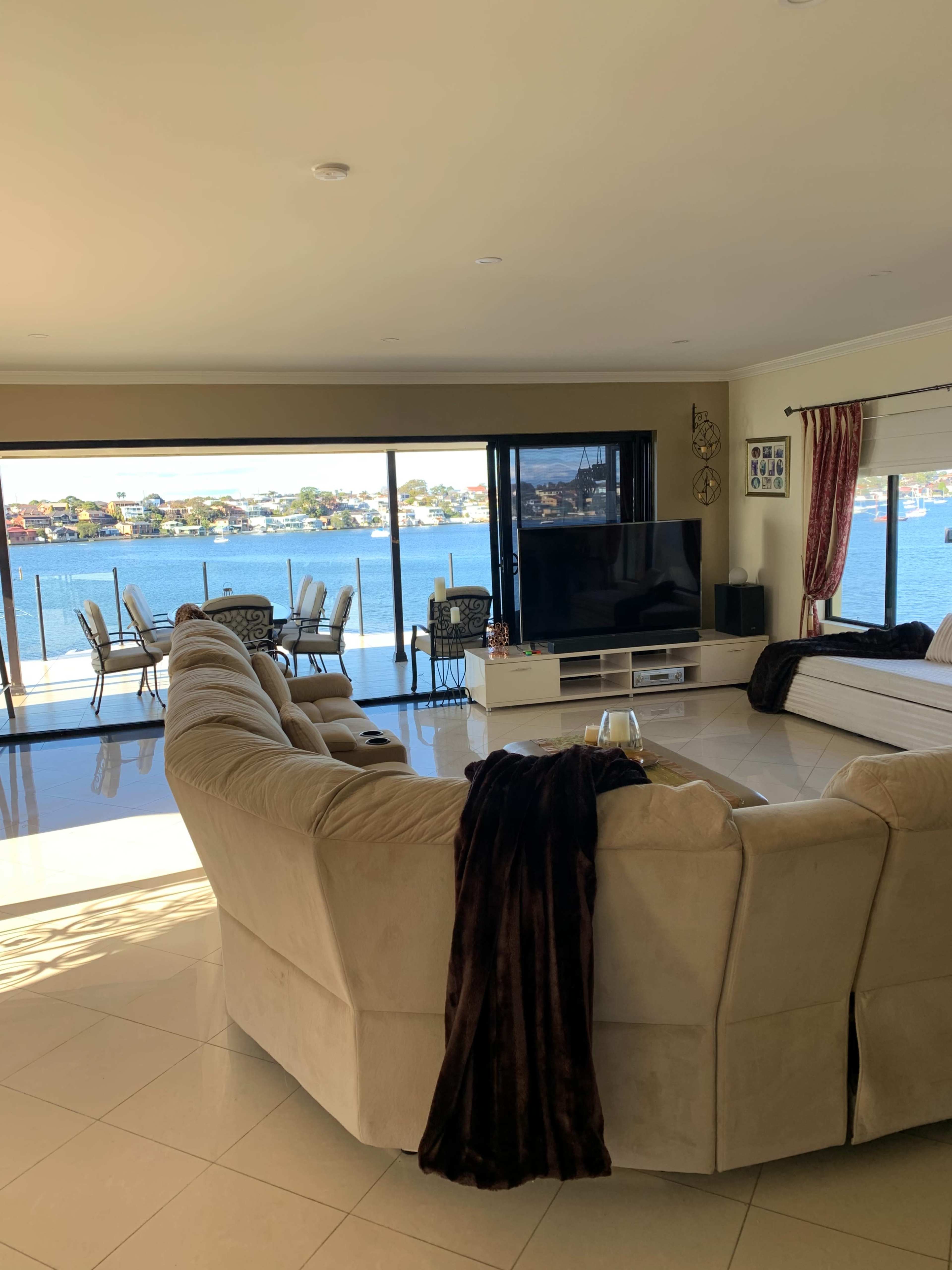A spacious living room features a large beige sofa, a TV console, and extensive windows overlooking a waterfront with outdoor seating.