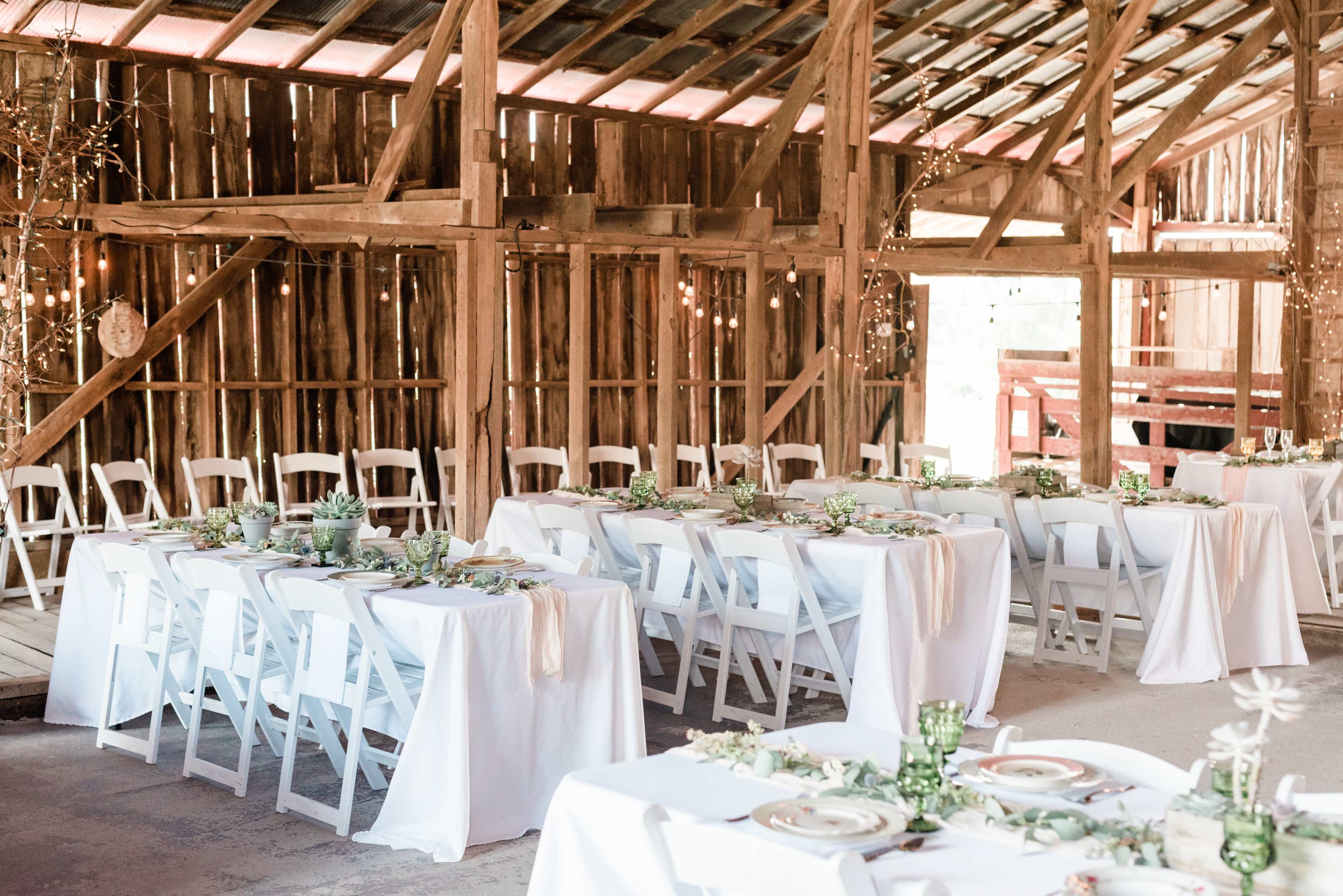 The image shows a rustic barn interior set up for a dining event, featuring tables with white tablecloths, plates, and greenery.