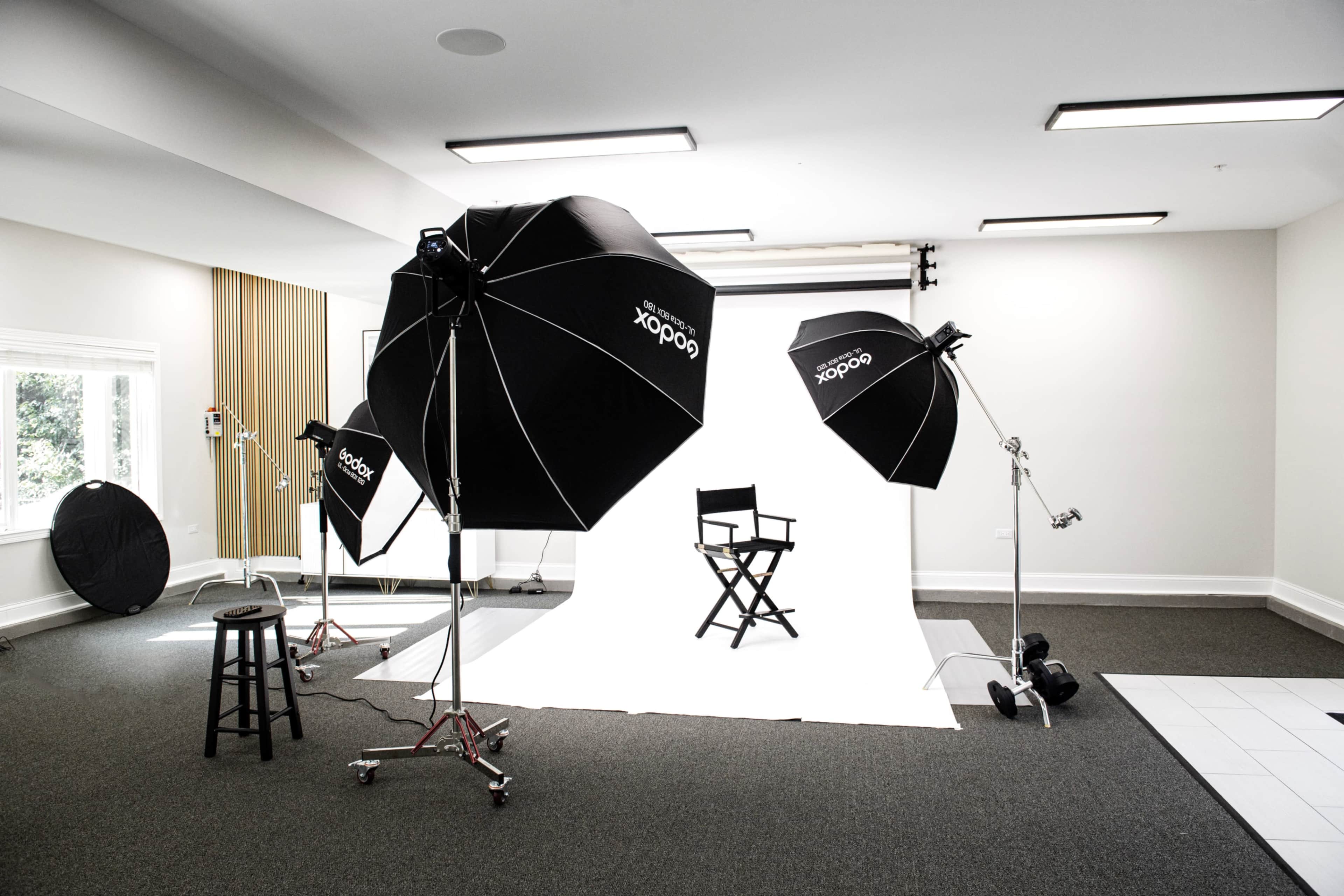A photography studio is set up with multiple light sources, a black director's chair, and a white backdrop.