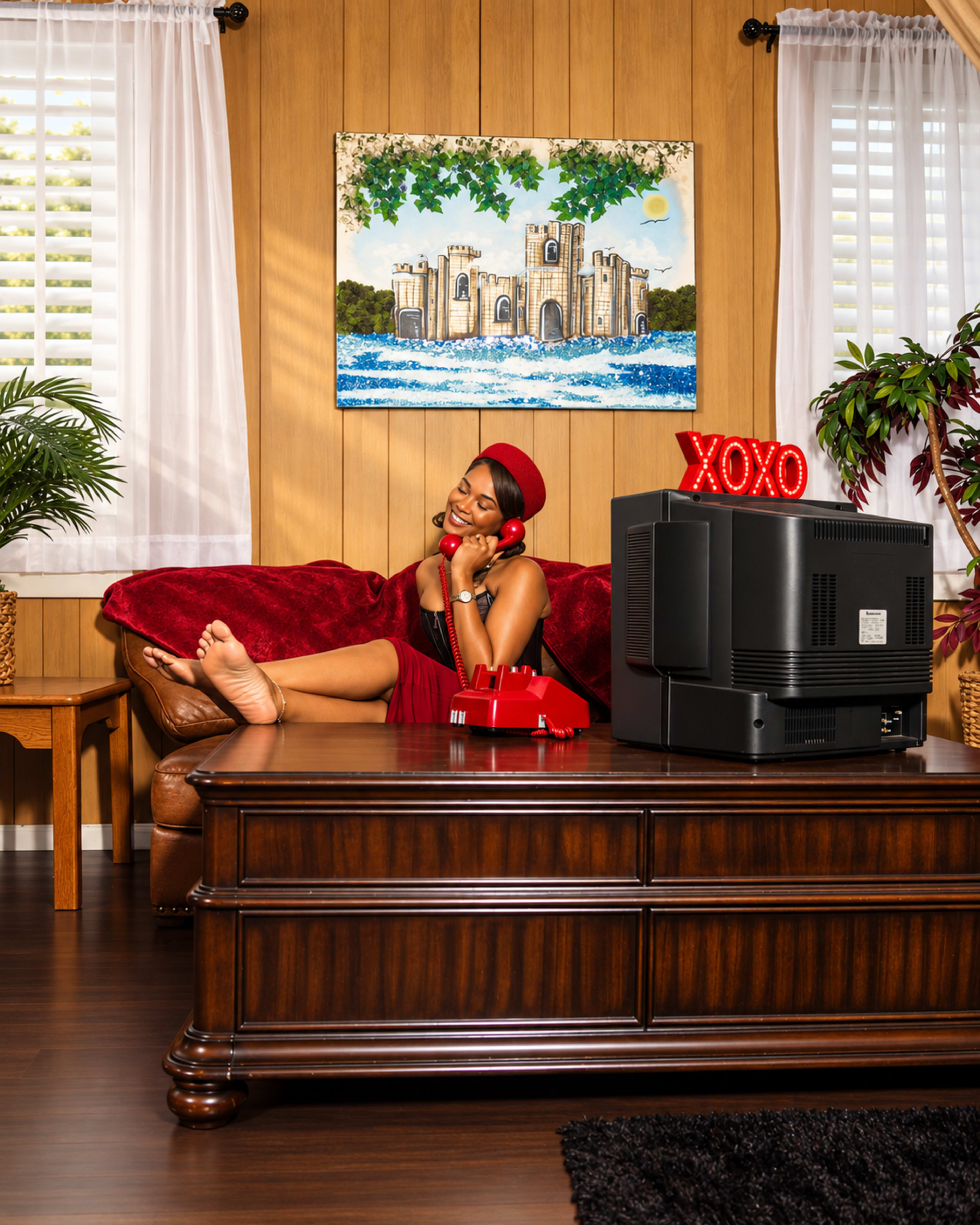 A woman in a red dress sits on a brown couch in a retro-styled living room, holding a red rotary phone beside a large TV displaying neon "XOXO" signage.