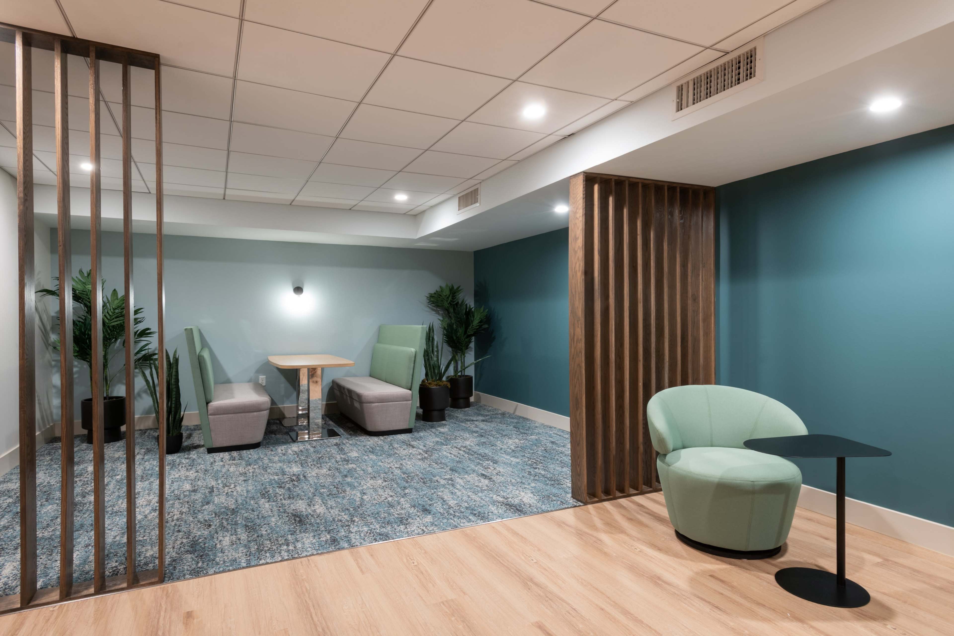 The image shows a modern lounge area featuring two upholstered chairs and a small table on a textured blue carpet, separated by wooden dividers.
