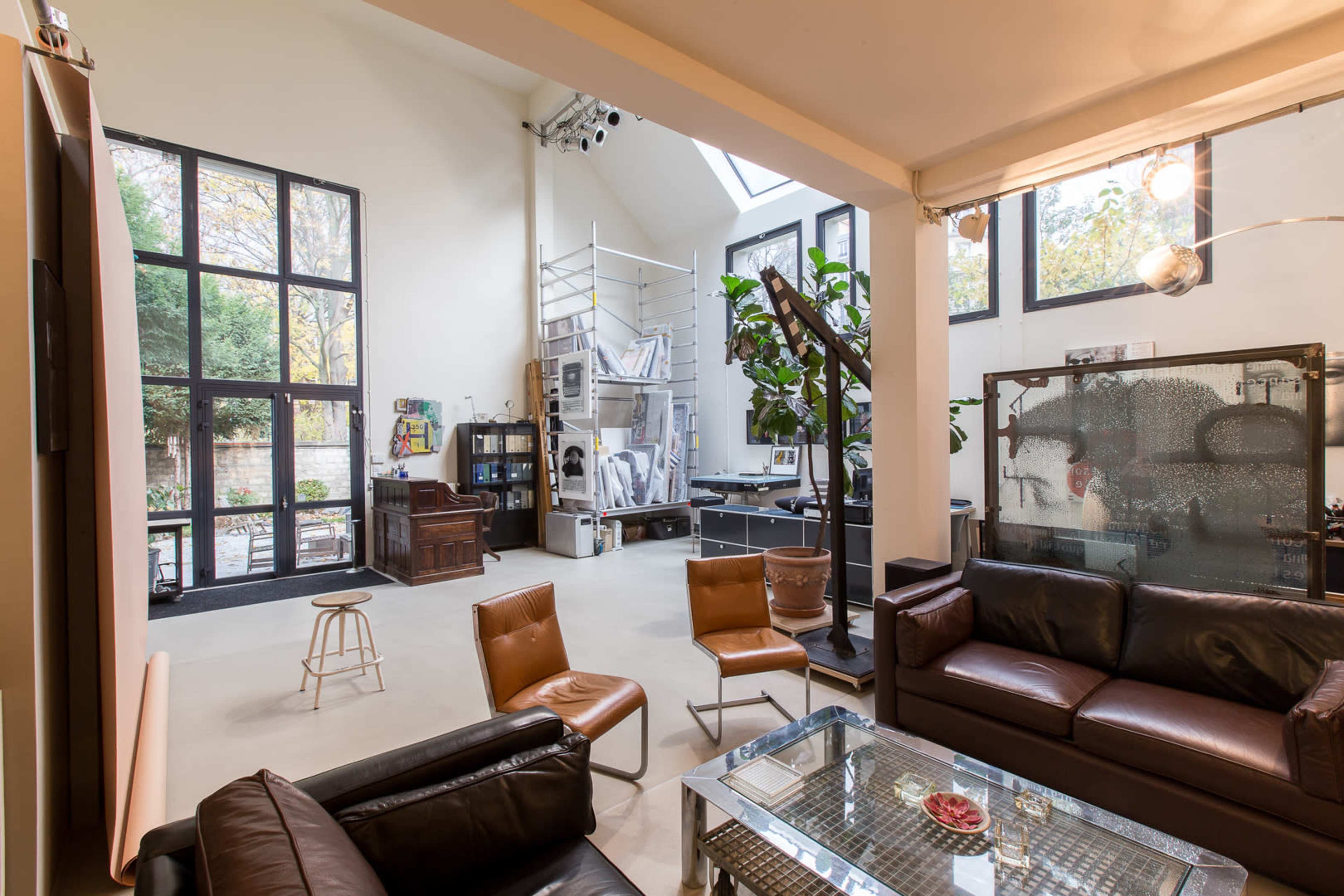 An open-concept living area featuring leather seating, a glass coffee table, large windows, and a workspace with a plant in the background.
