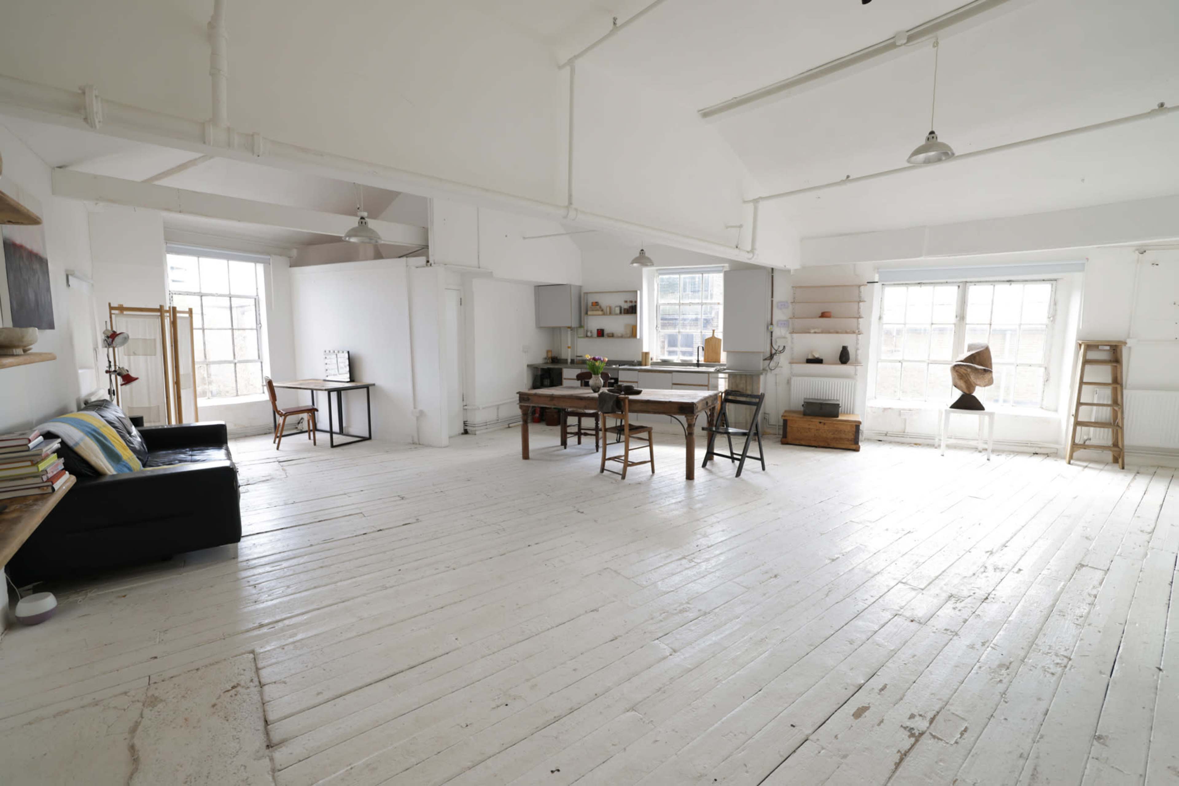 The image depicts a spacious, minimalist interior of a bright, white-walled room with wooden flooring, featuring a seating area, dining table, and various decor elements.