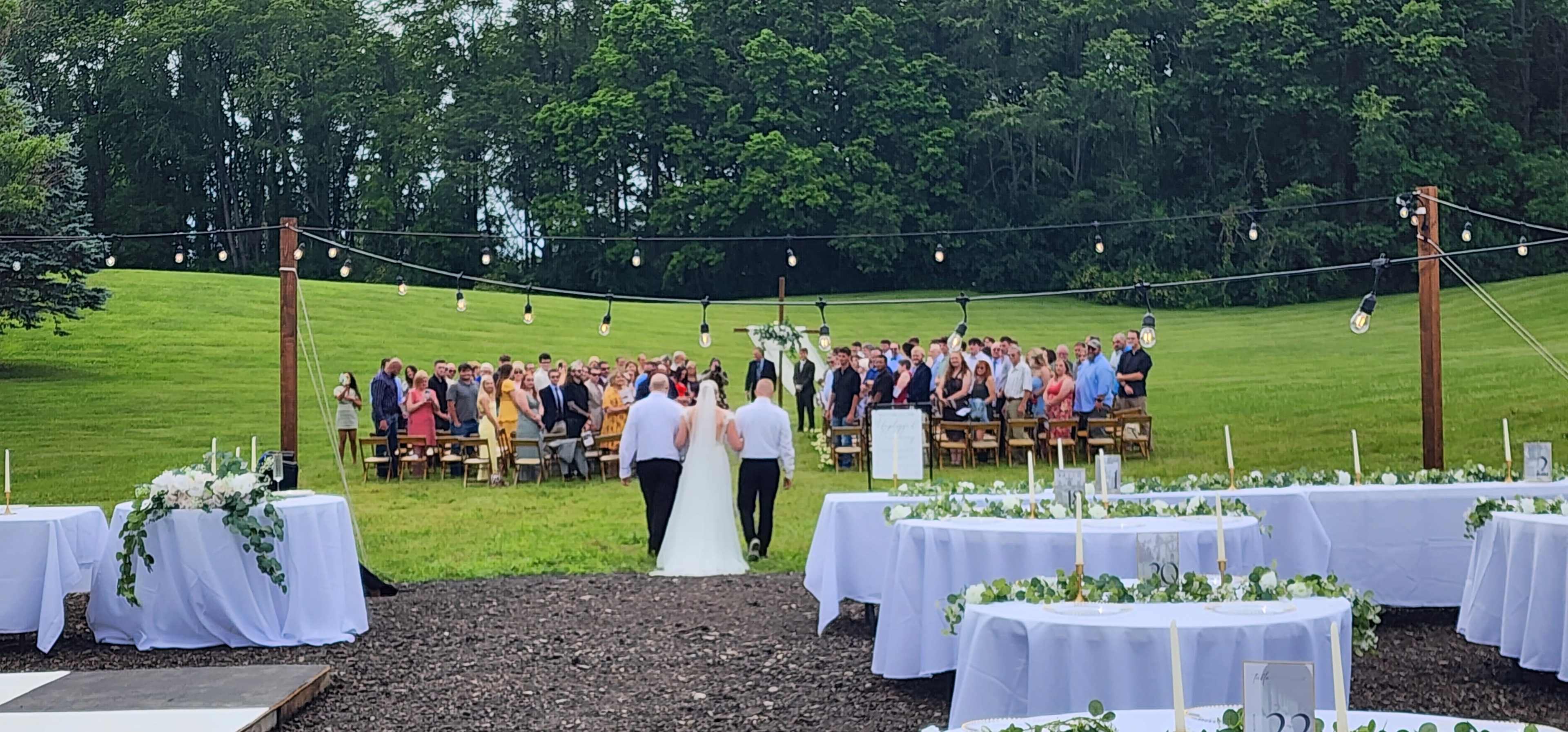 A couple walks down an aisle toward a gathering of guests seated on benches in an outdoor wedding ceremony setting surrounded by greenery.