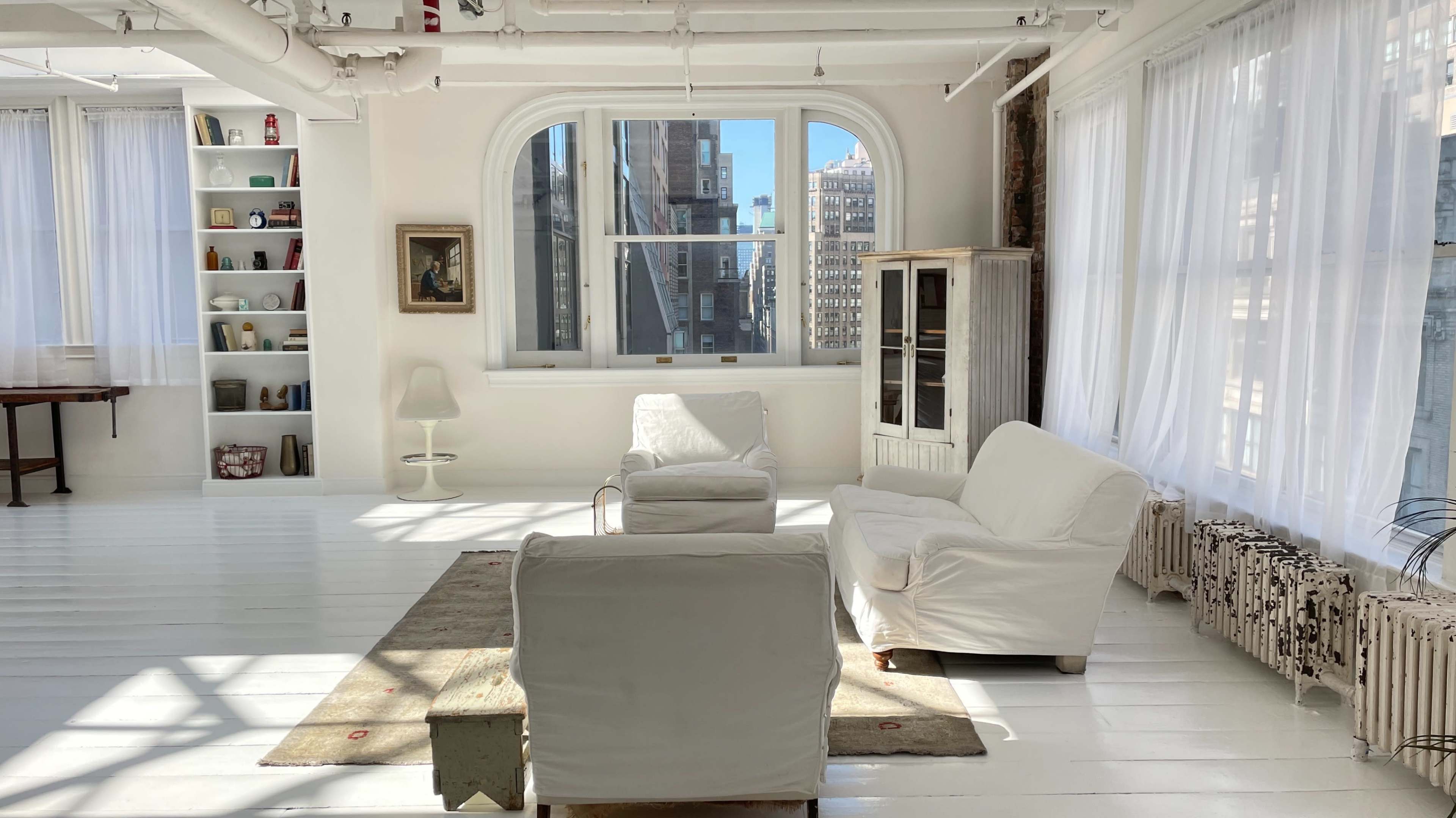 A bright, airy room with large windows, white walls, a rug on the wooden floor, and two white upholstered chairs facing a pale wooden cabinet.