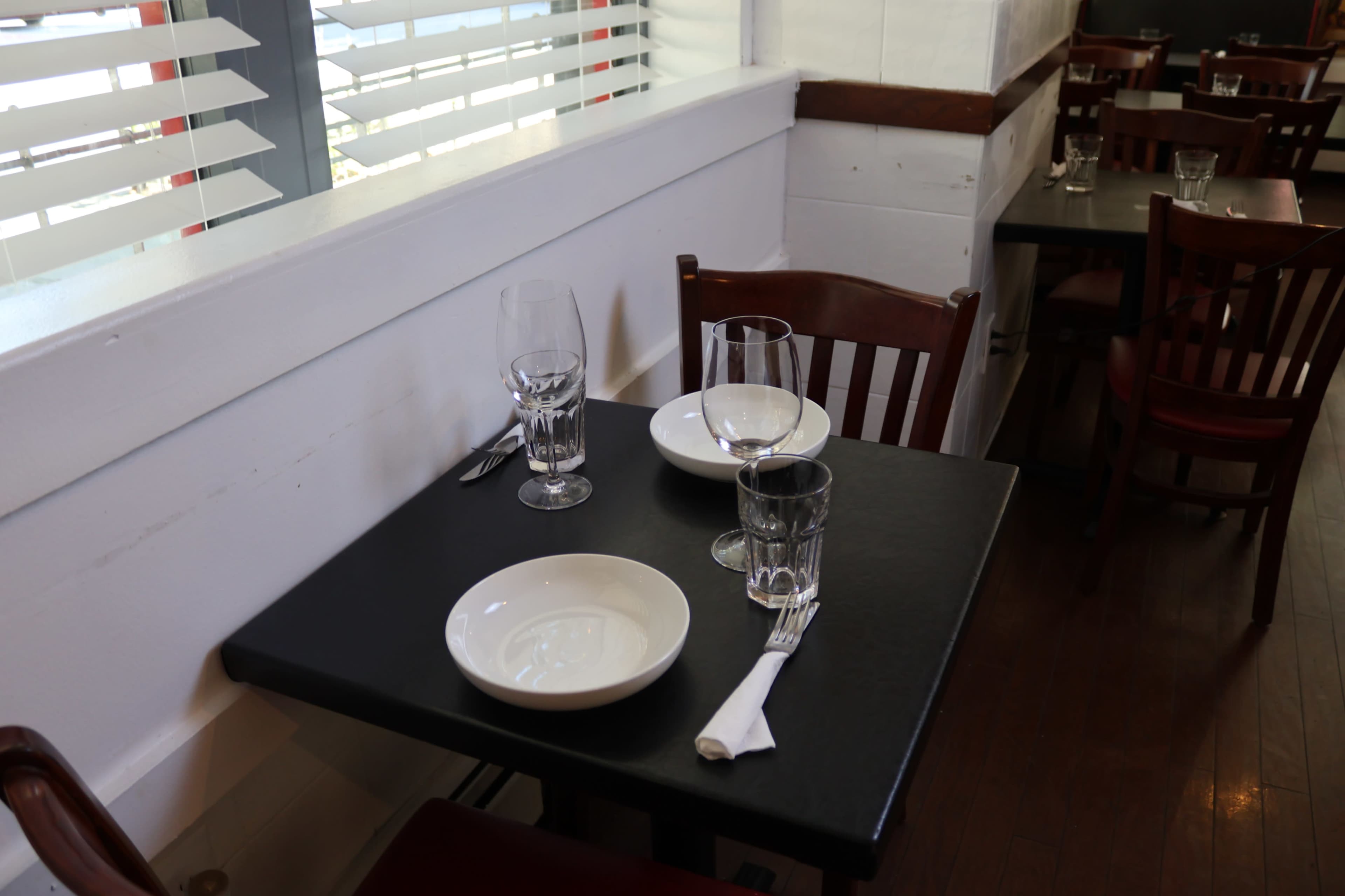 A table set for dining with empty plates, glasses, and utensils is positioned in a restaurant with wooden chairs and white walls.