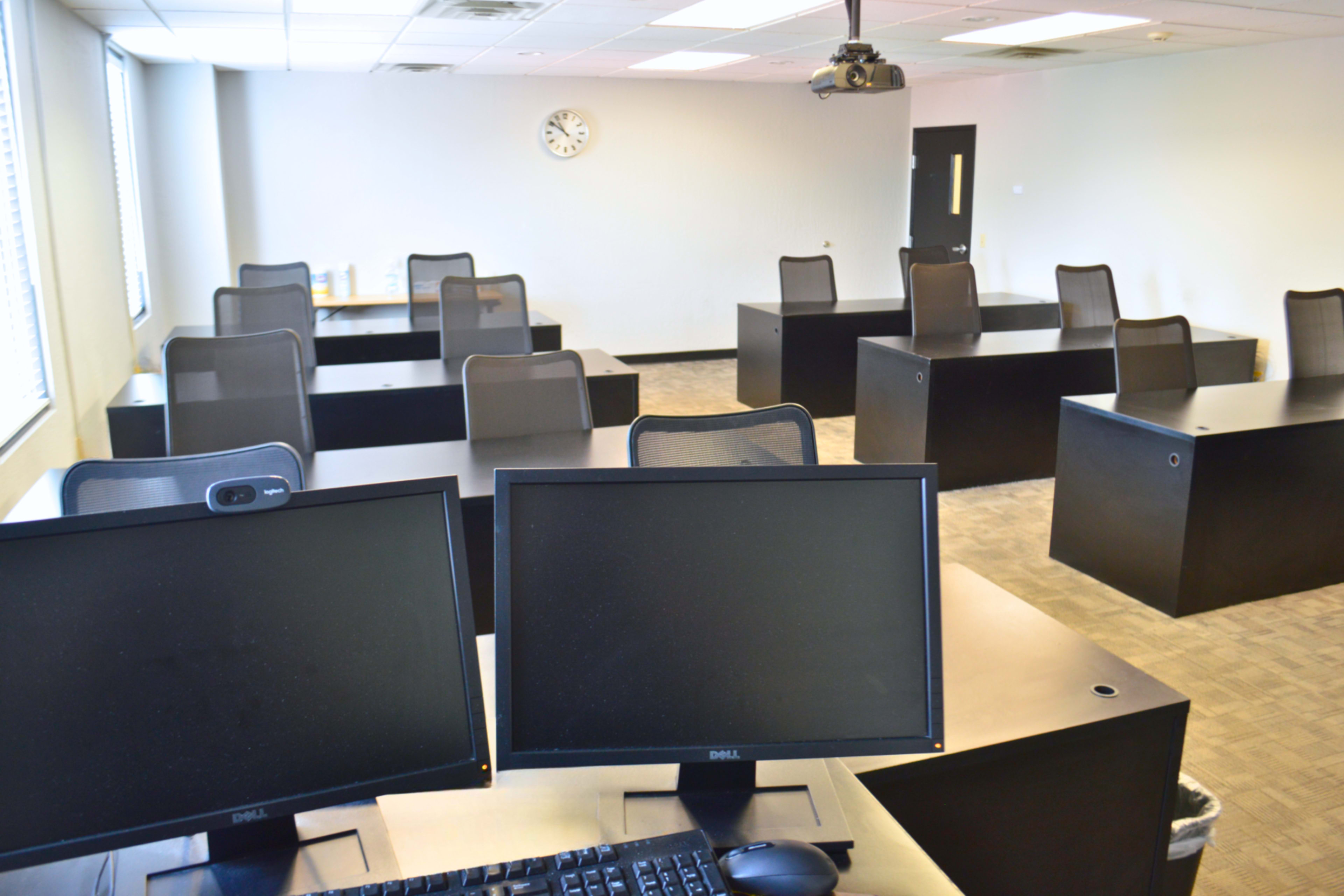 The image shows a modern classroom with several desks and computers arranged in rows and a clock on the wall.