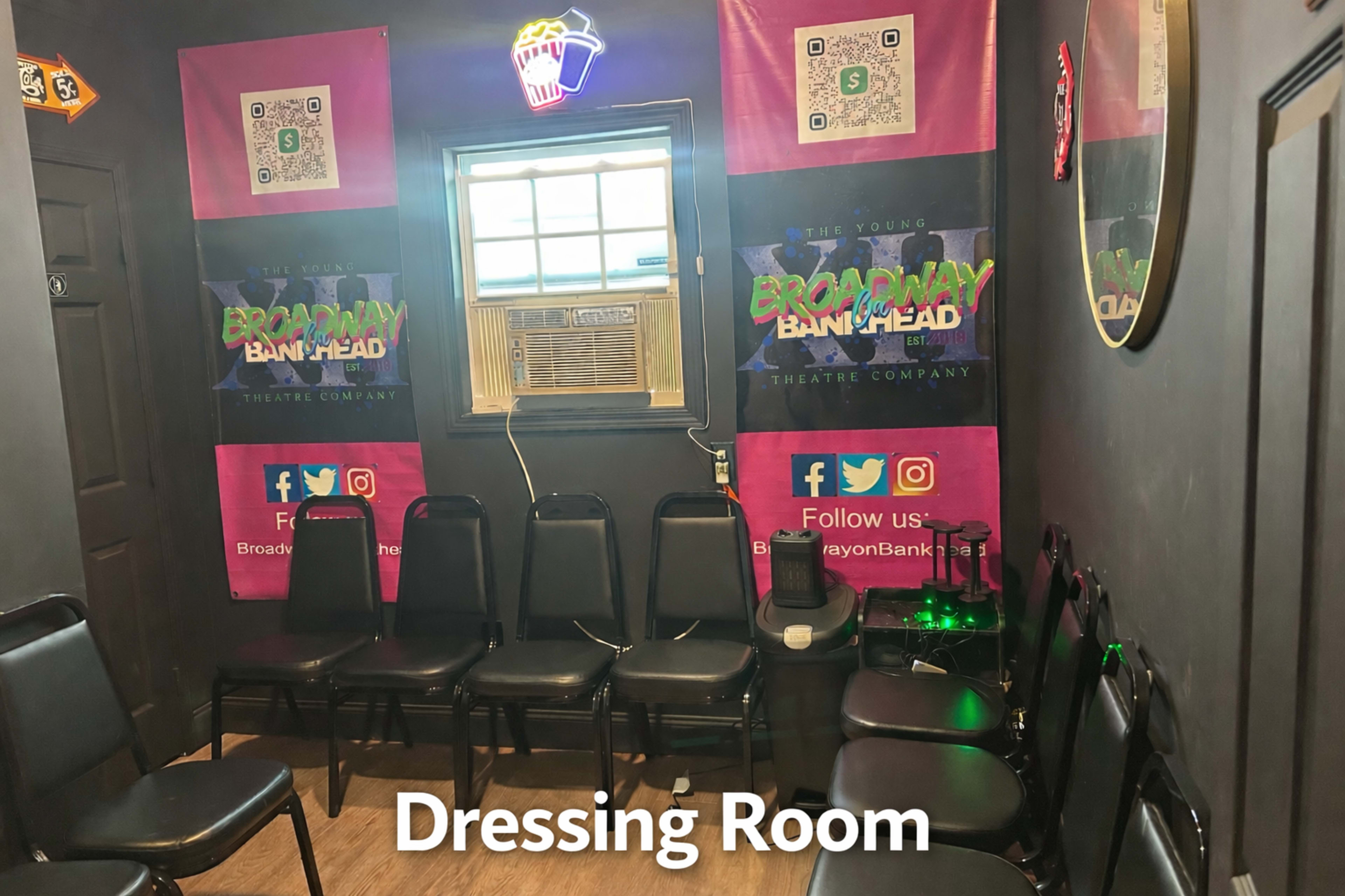 The image shows a dimly lit dressing room with black walls, several black chairs arranged in a row, and colorful posters on the walls promoting a theatre company.