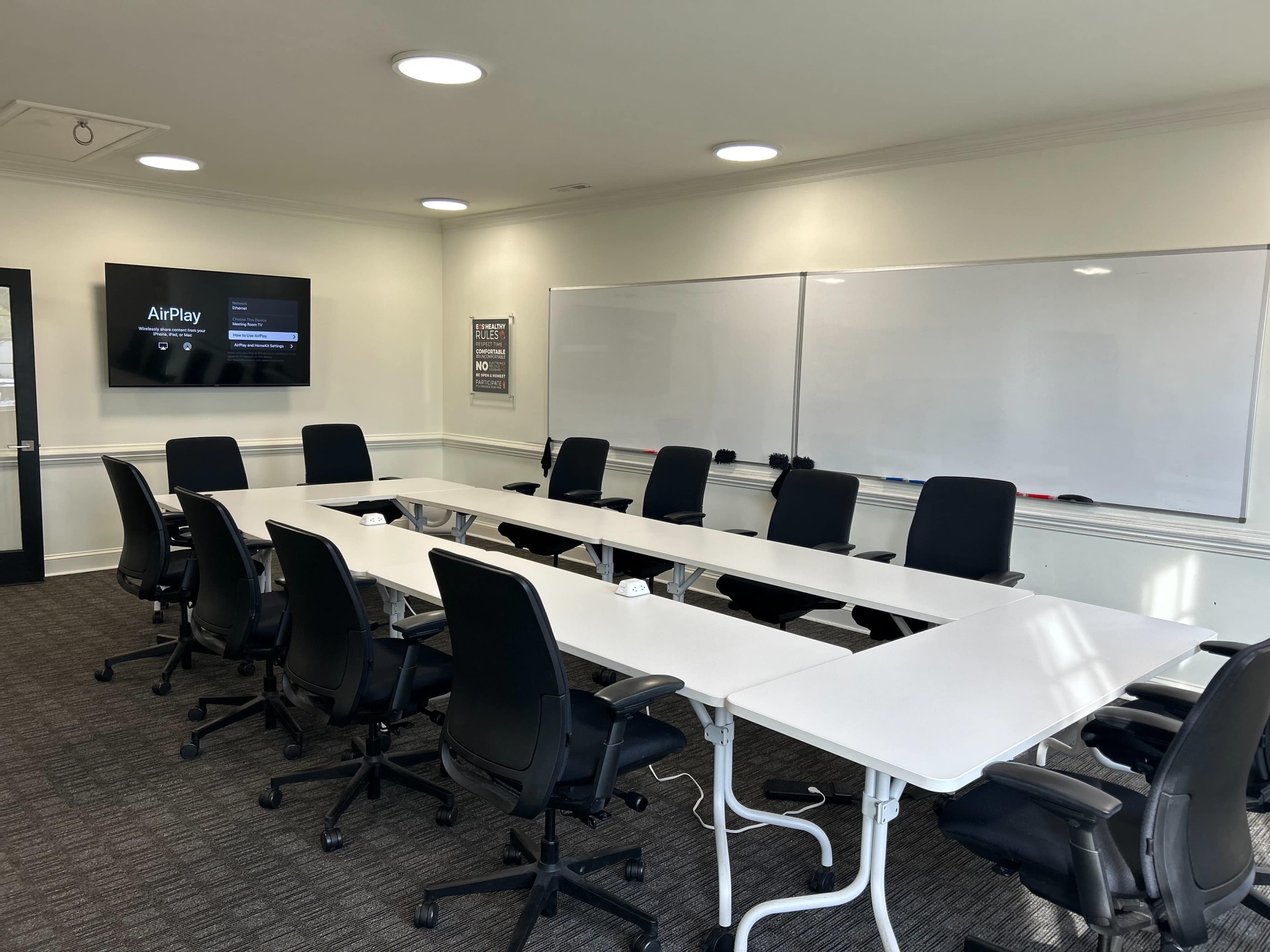 A conference room is equipped with a long table, multiple black office chairs, and a screen displaying "AirPlay" on one wall.