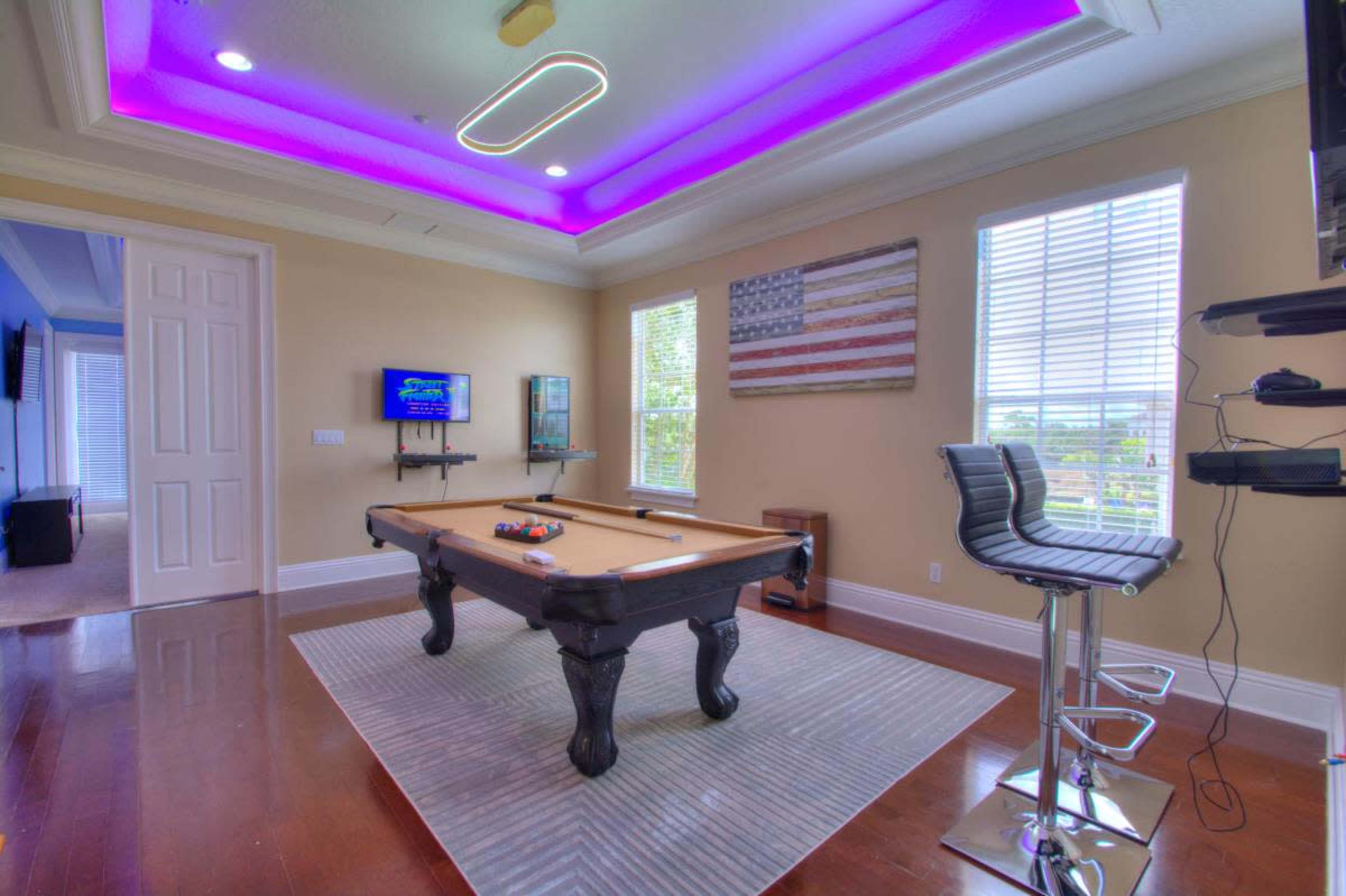 The image shows a well-lit room featuring a billiards table, two bar stools, and an American flag on the wall.