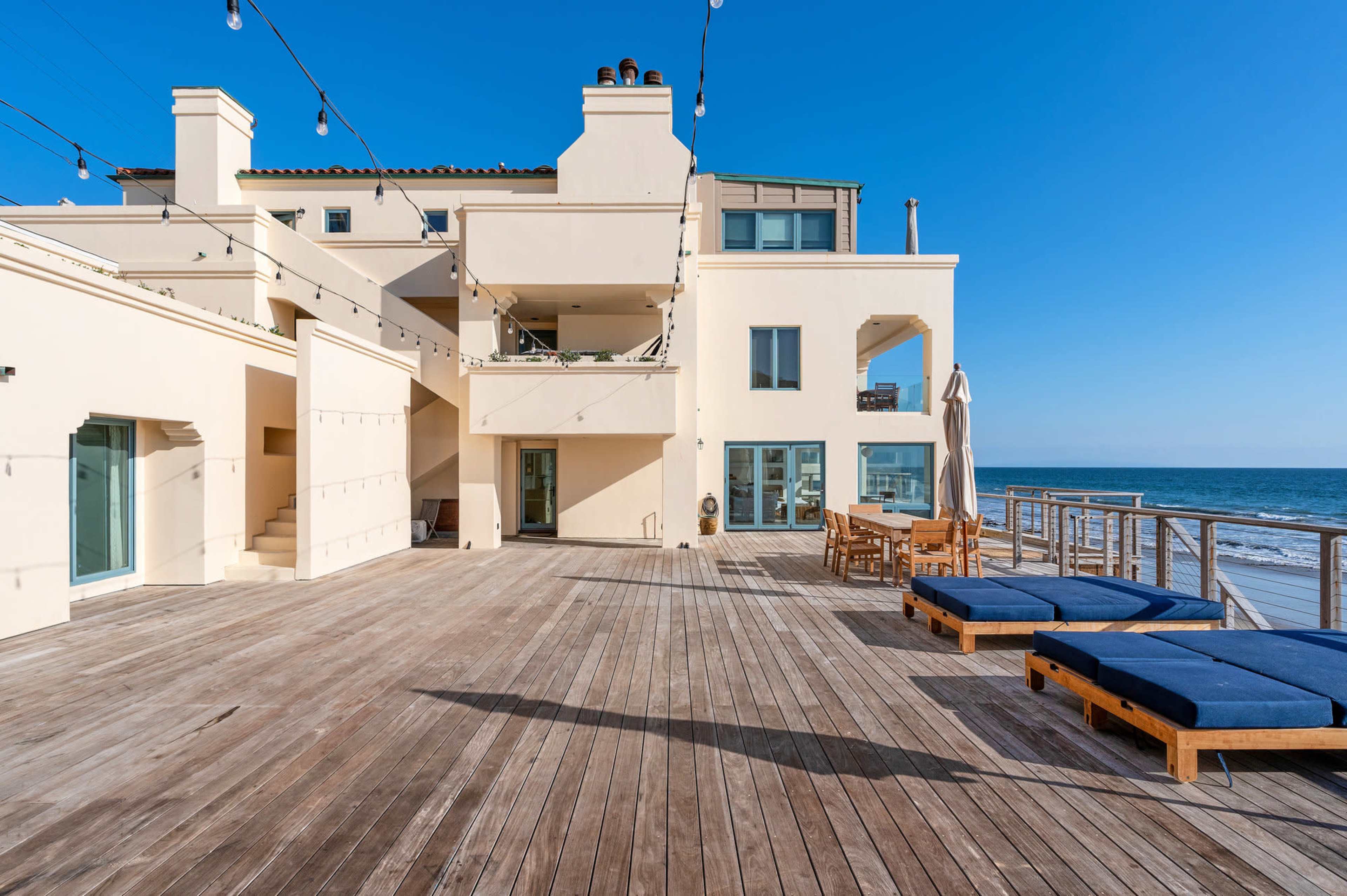 A modern coastal house with a wooden deck and lounge chairs overlooks the ocean under a clear blue sky.