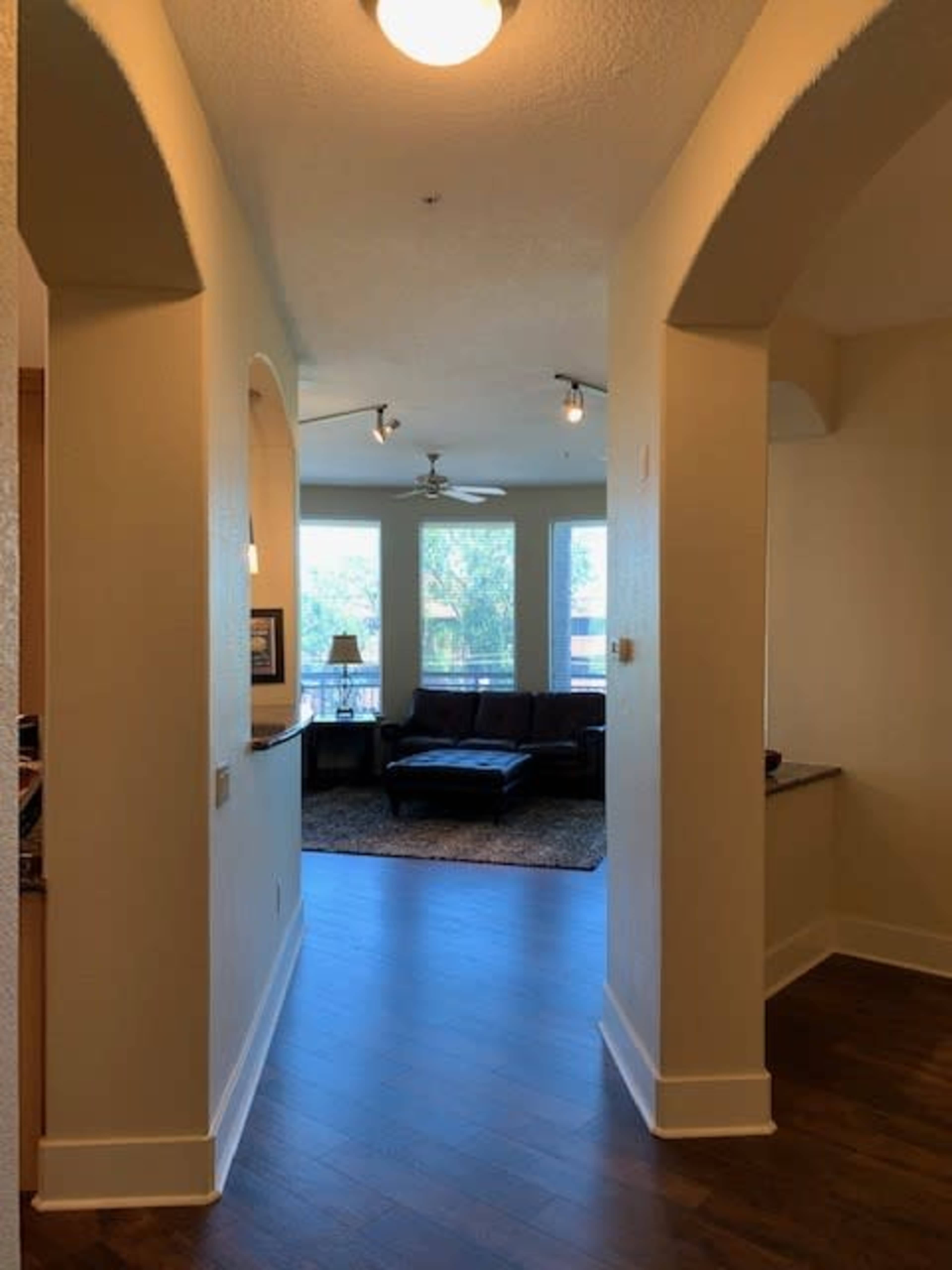 The image shows a hallway leading to a living room with large windows and a leather couch.