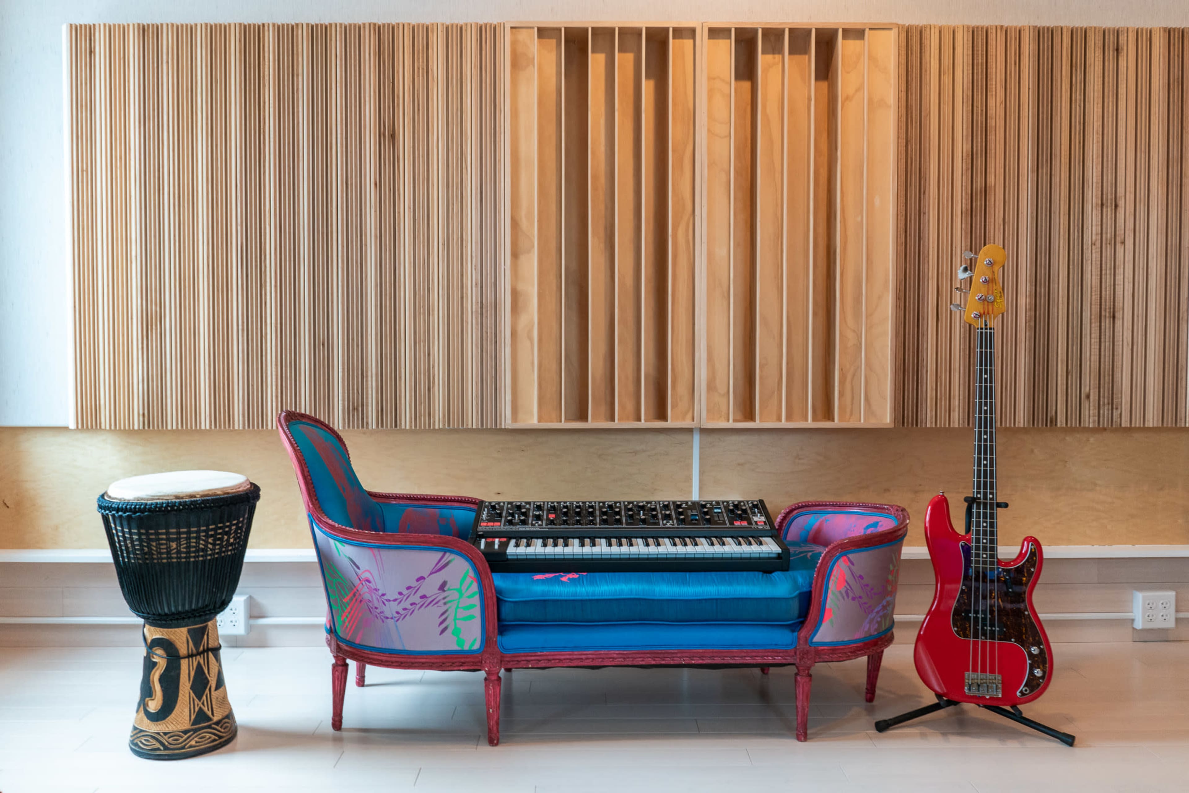 A colorful couch holds a keyboard surrounded by a drum and a bass guitar against a backdrop of wooden acoustic panels.