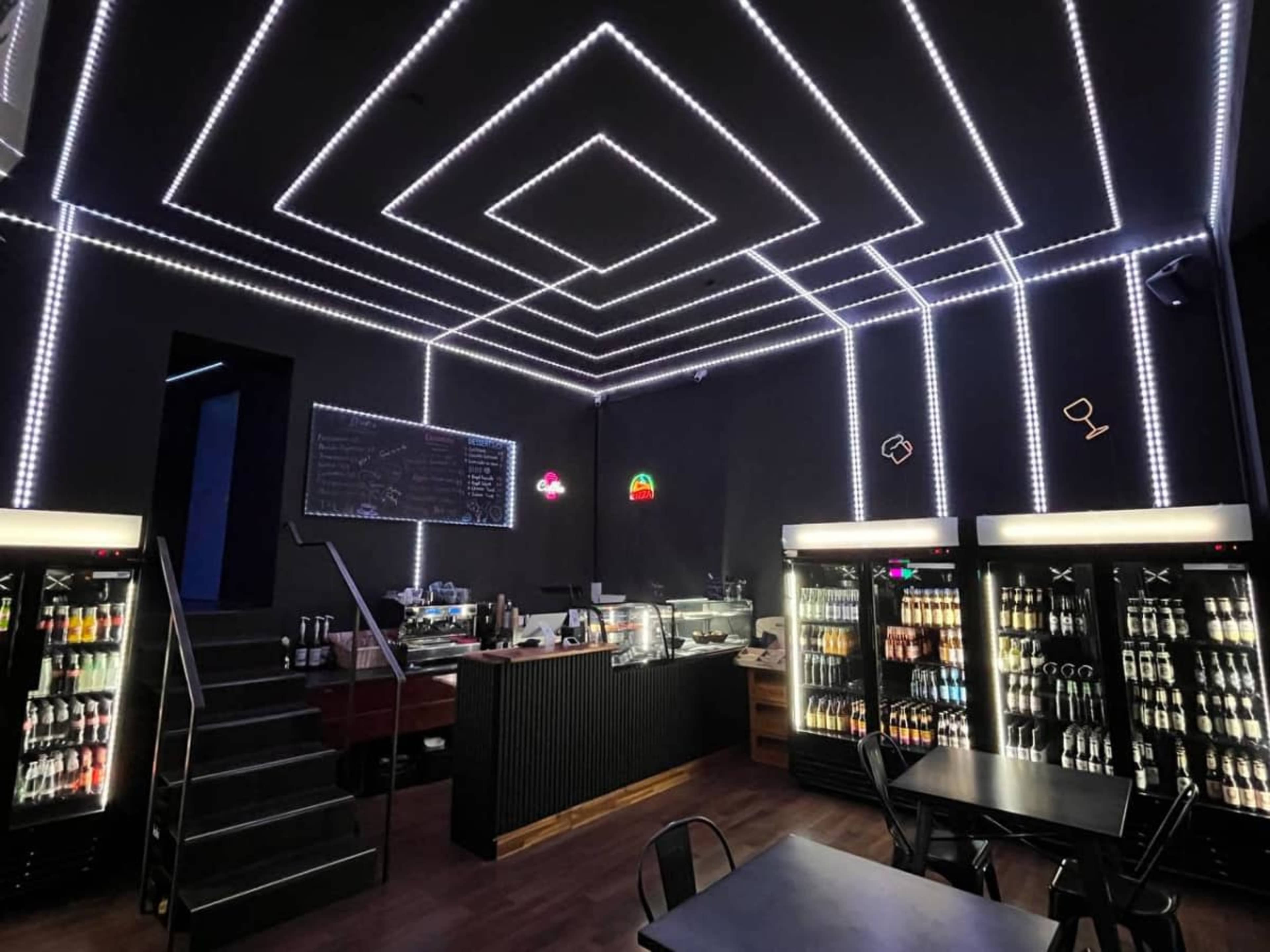 The interior of a modern cafe features a black ceiling with LED strip lights creating geometric patterns, glass display refrigerators stocked with drinks, and a counter with coffee equipment.