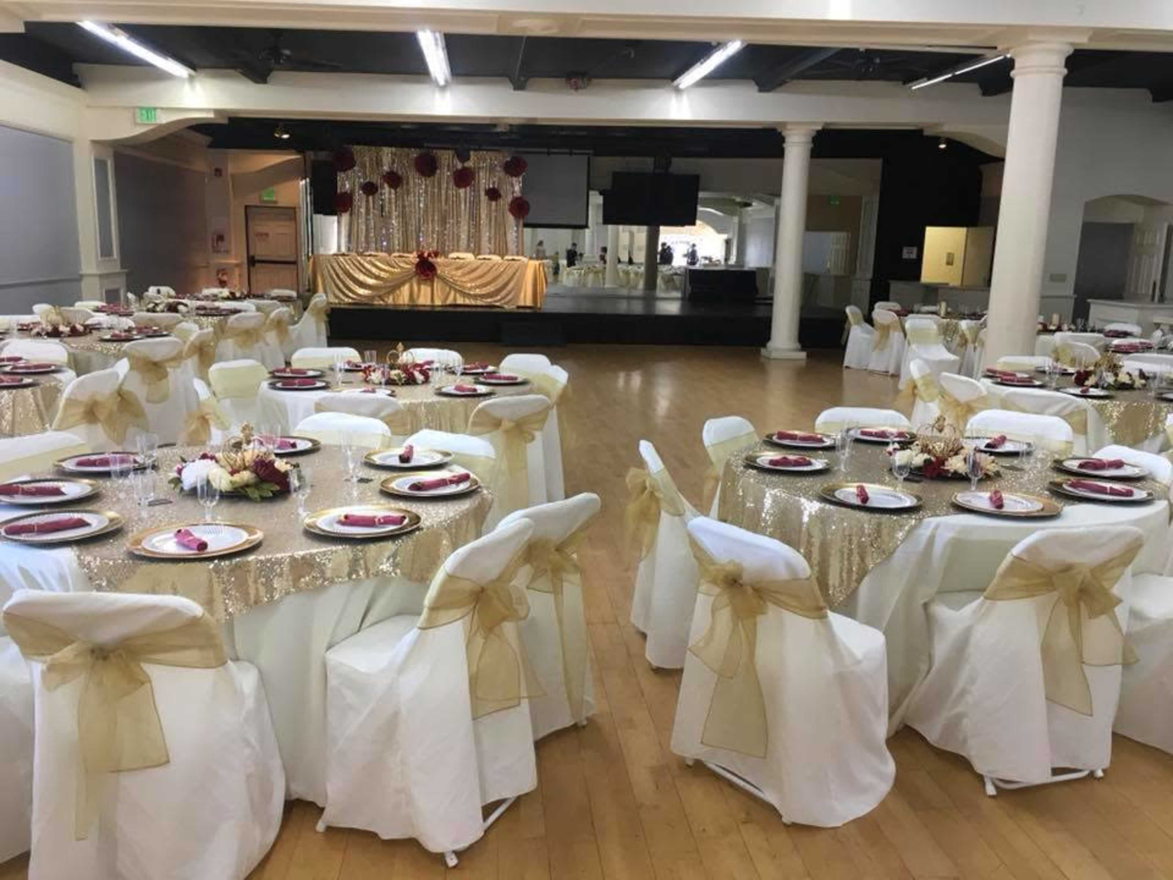 The image shows a banquet hall set up for an event, with multiple round tables covered in gold sequined tablecloths, each adorned with white floral centerpieces and gold-accented place settings.